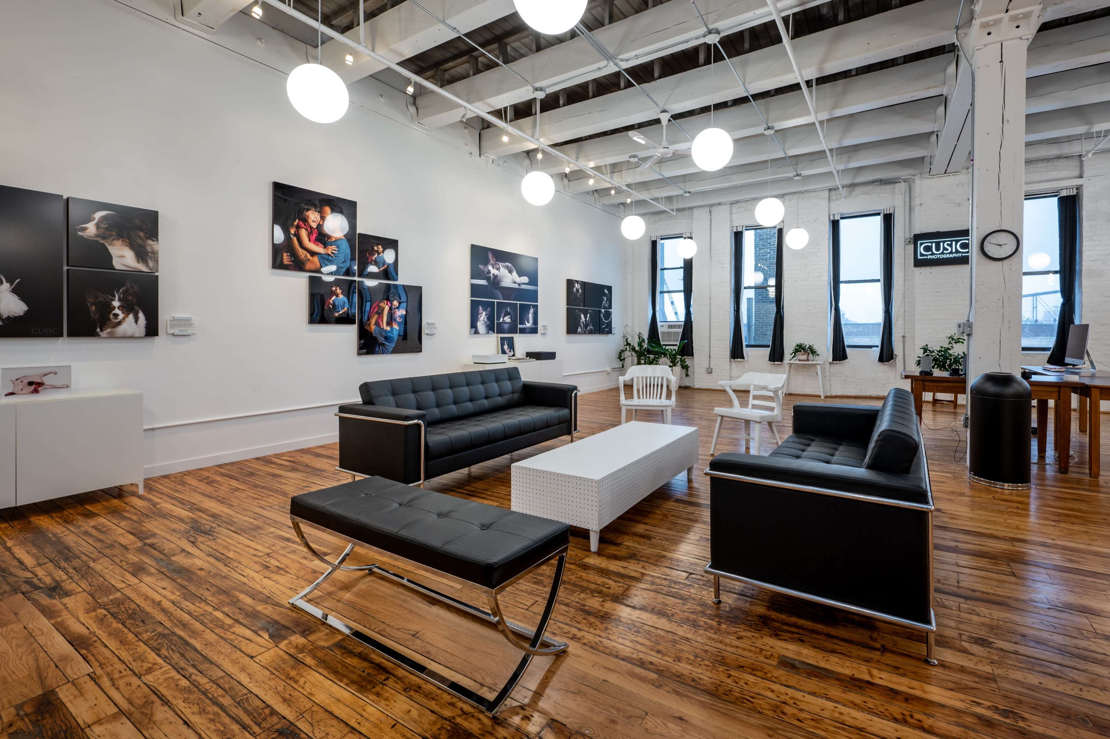 A modern interior space with black leather sofas, a white coffee table, and several framed photographs on the white walls, illuminated by hanging round light fixtures.