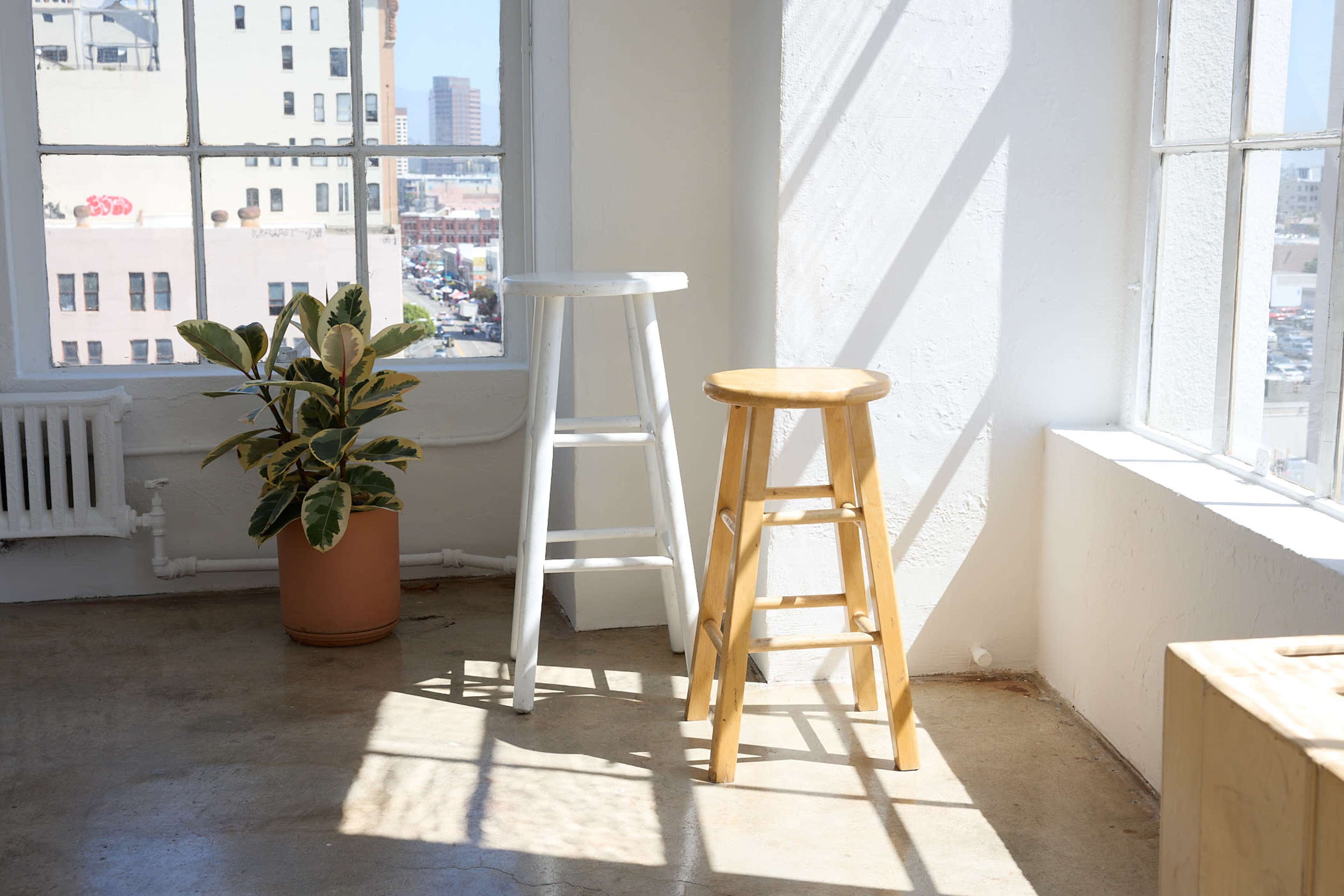 Two wooden stools, one white and one natural wood, positioned near a window with a plant in a pot beside them, casting shadows on the concrete floor.
