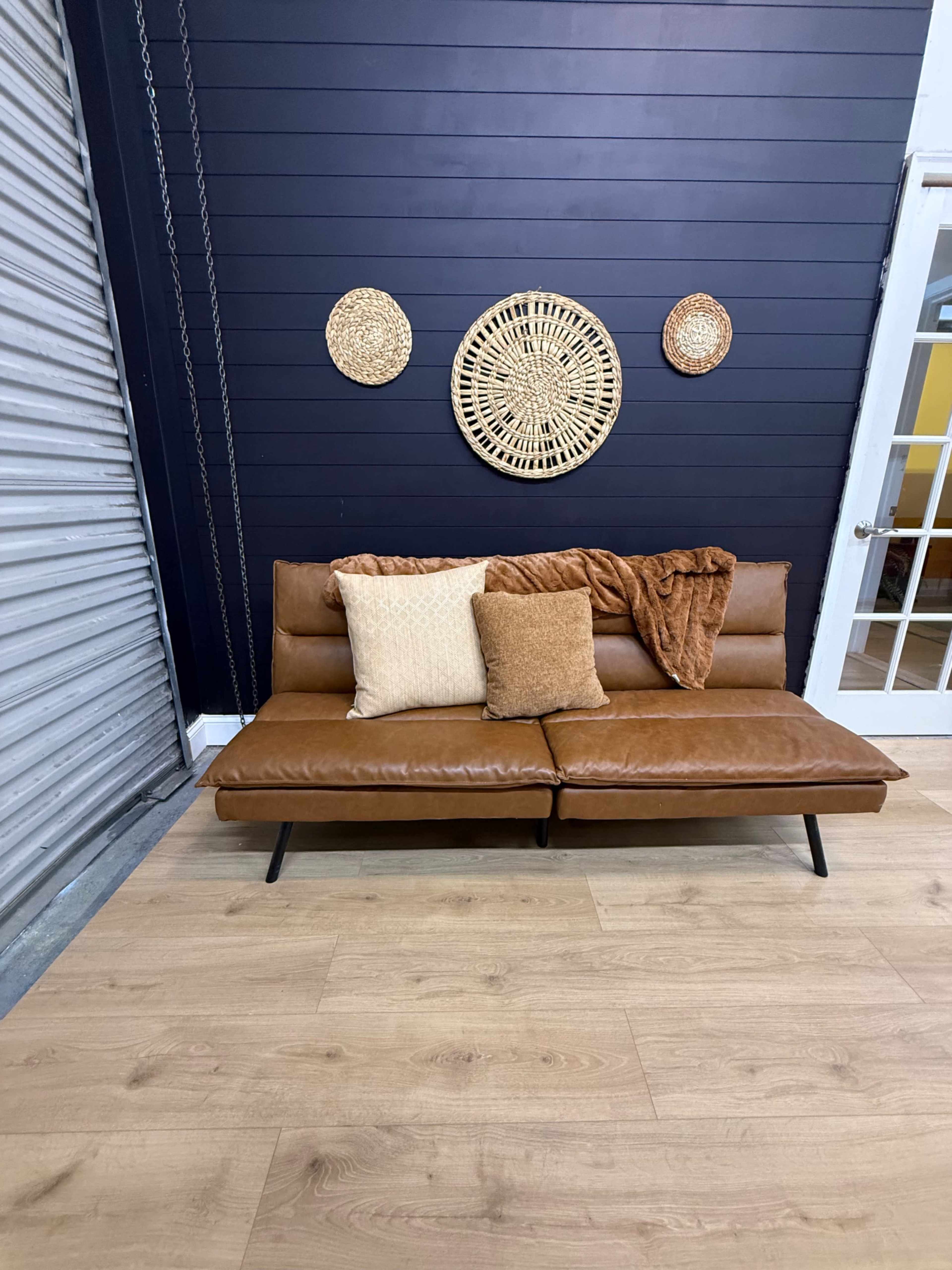 The image features a brown leather sofa with two pillows and a blanket, situated against a dark blue wall adorned with three circular woven decorations.