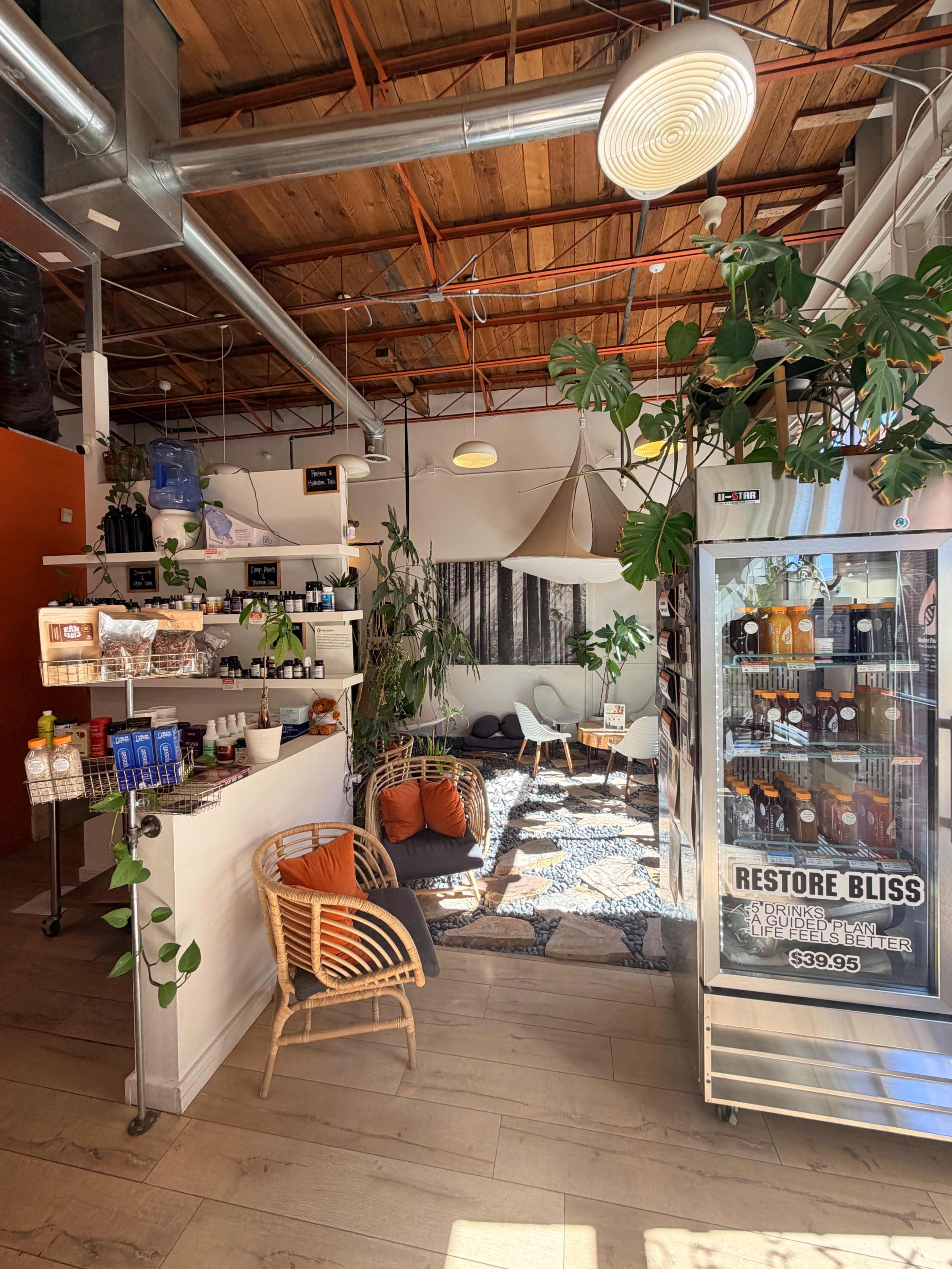 The image shows a cozy wellness spa interior featuring a reception area with a service counter, plants, seating, and a glass refrigerator displaying products.