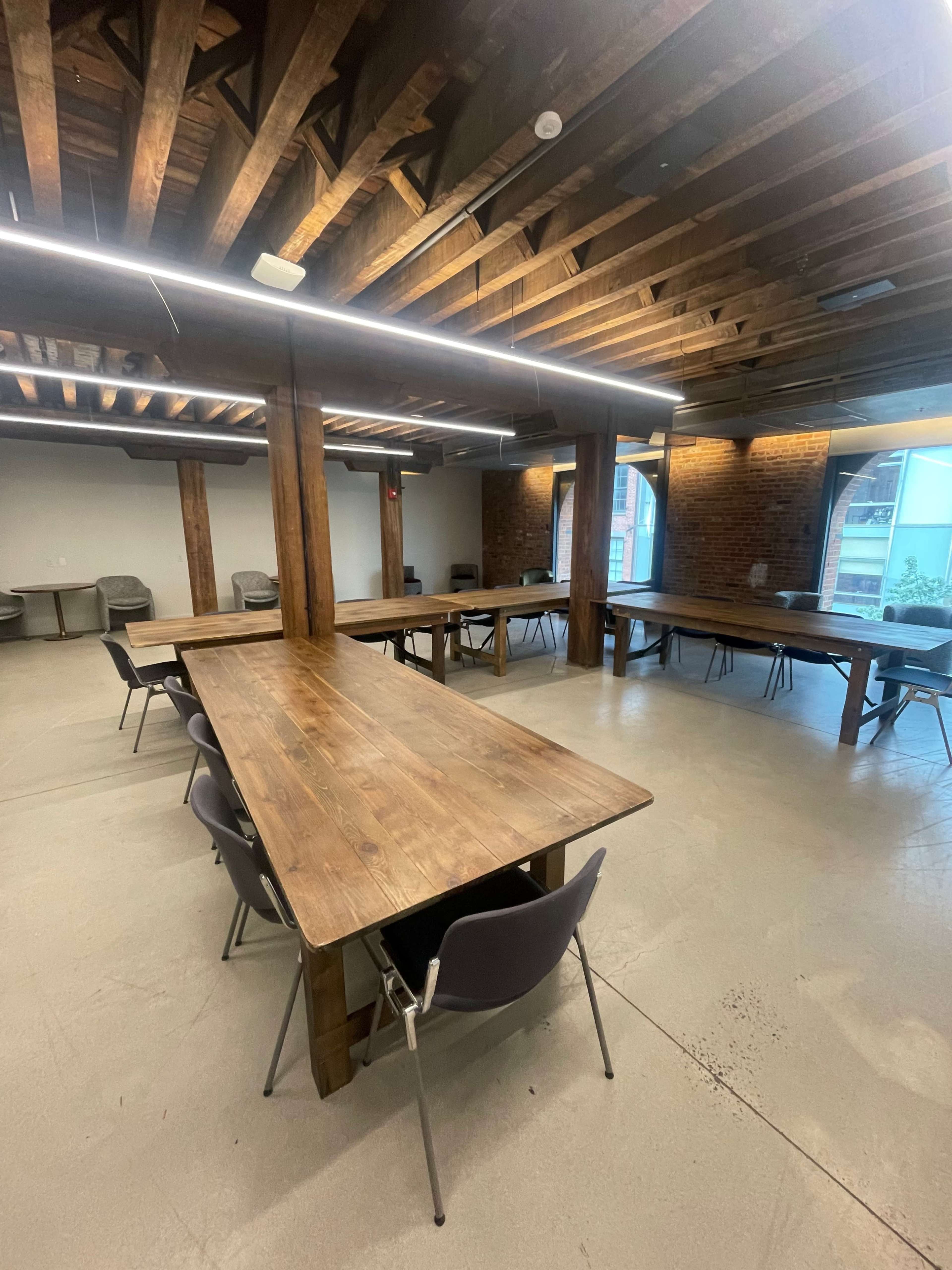 A spacious meeting room with wooden beams and tables arranged in a workshop layout, surrounded by chairs.