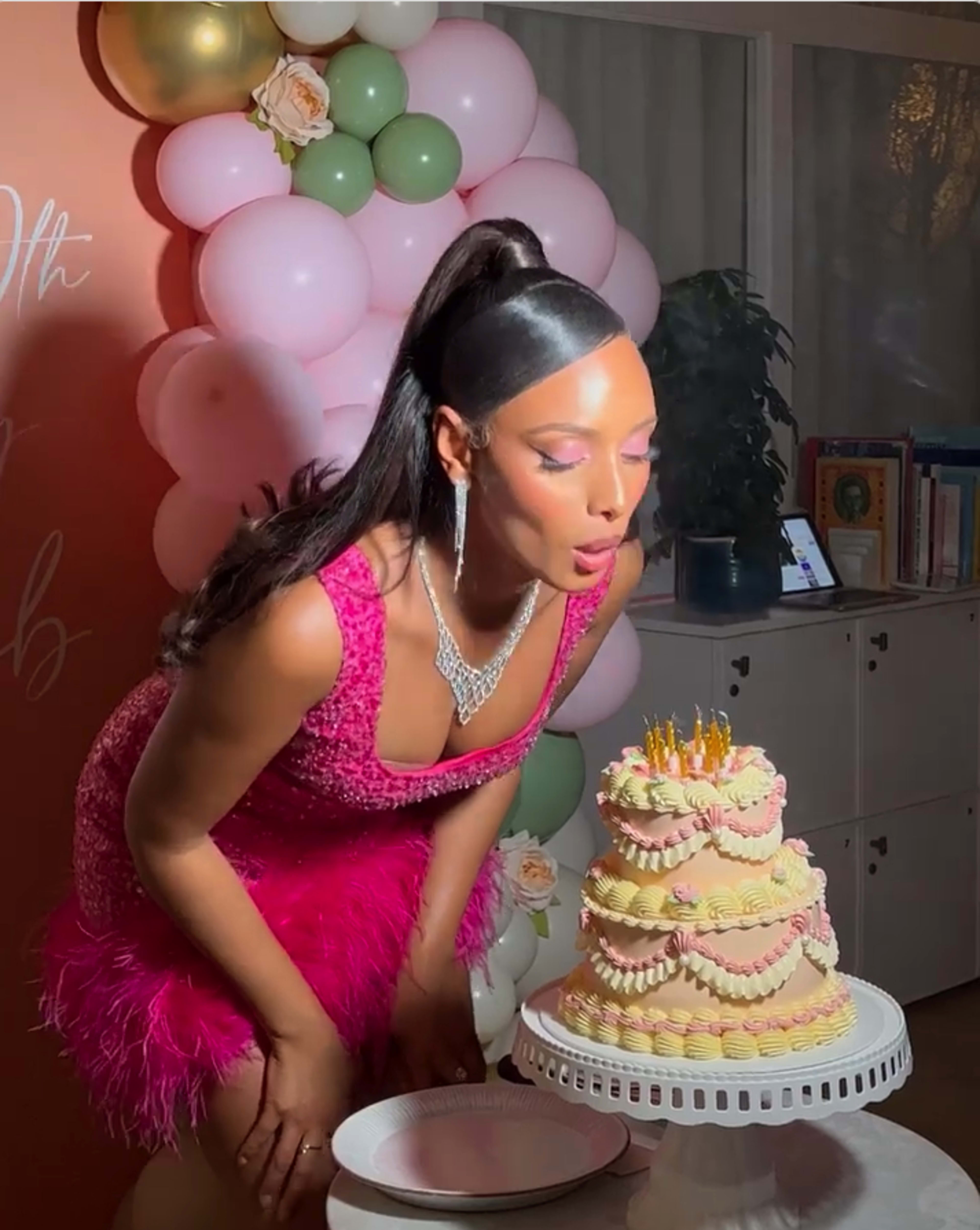 A woman in a pink dress is blowing out candles on a multi-tiered birthday cake, surrounded by balloons and decorations.