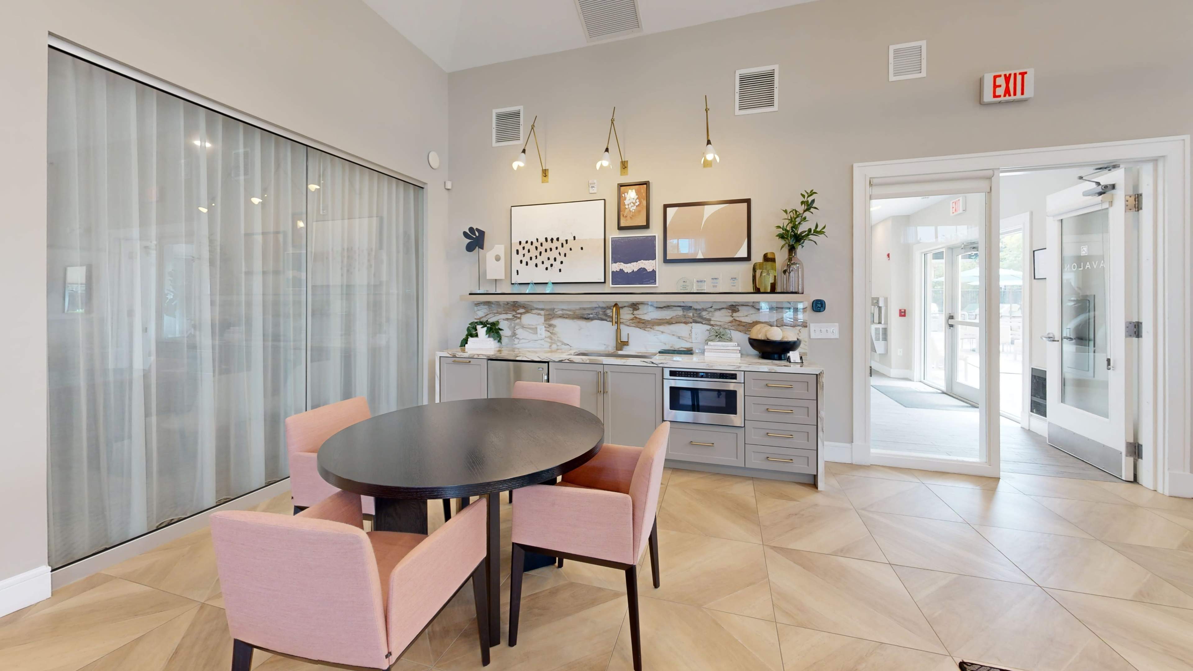 A modern kitchen area features a round black table surrounded by four pink upholstered chairs, with a countertop, microwave, and artwork on the wall.