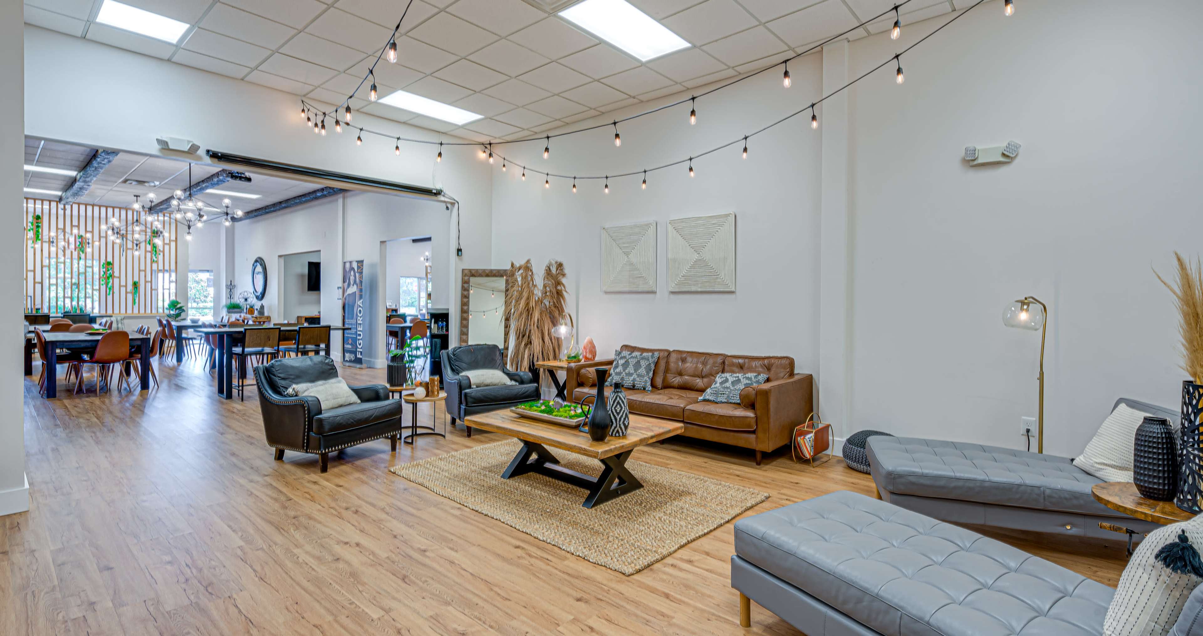 A modern lounge area with a brown leather sofa, two lounge chairs, and a coffee table on a wooden floor, surrounded by string lights and an open layout leading to a dining area.