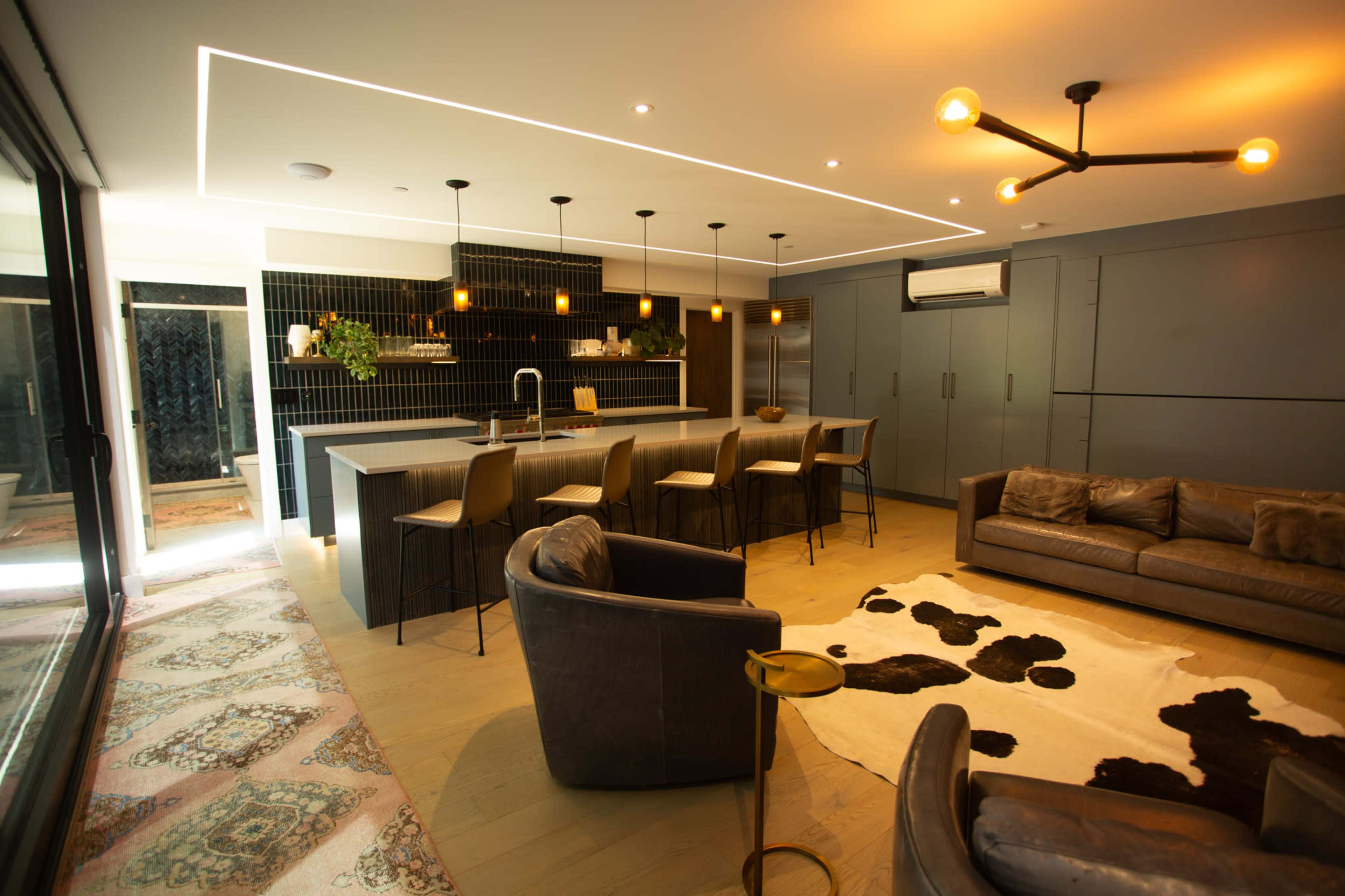 The image shows a modern kitchen and living area featuring a large island with bar stools, stylish lighting fixtures, and a sliding glass door leading to an outdoor space.