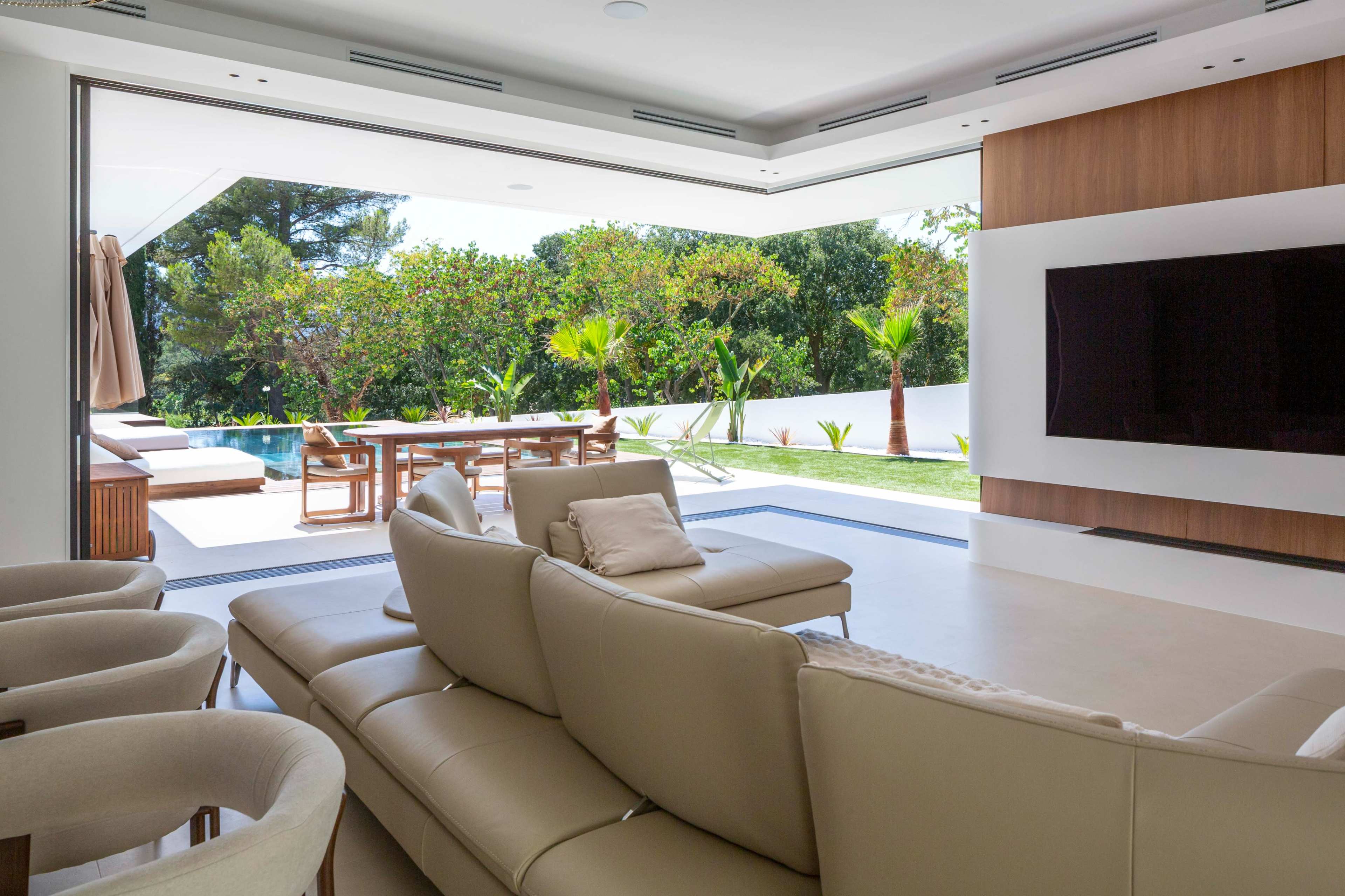 The image shows a modern living room with a light-colored sofa, large glass doors opening to a backyard with a pool and lush greenery.