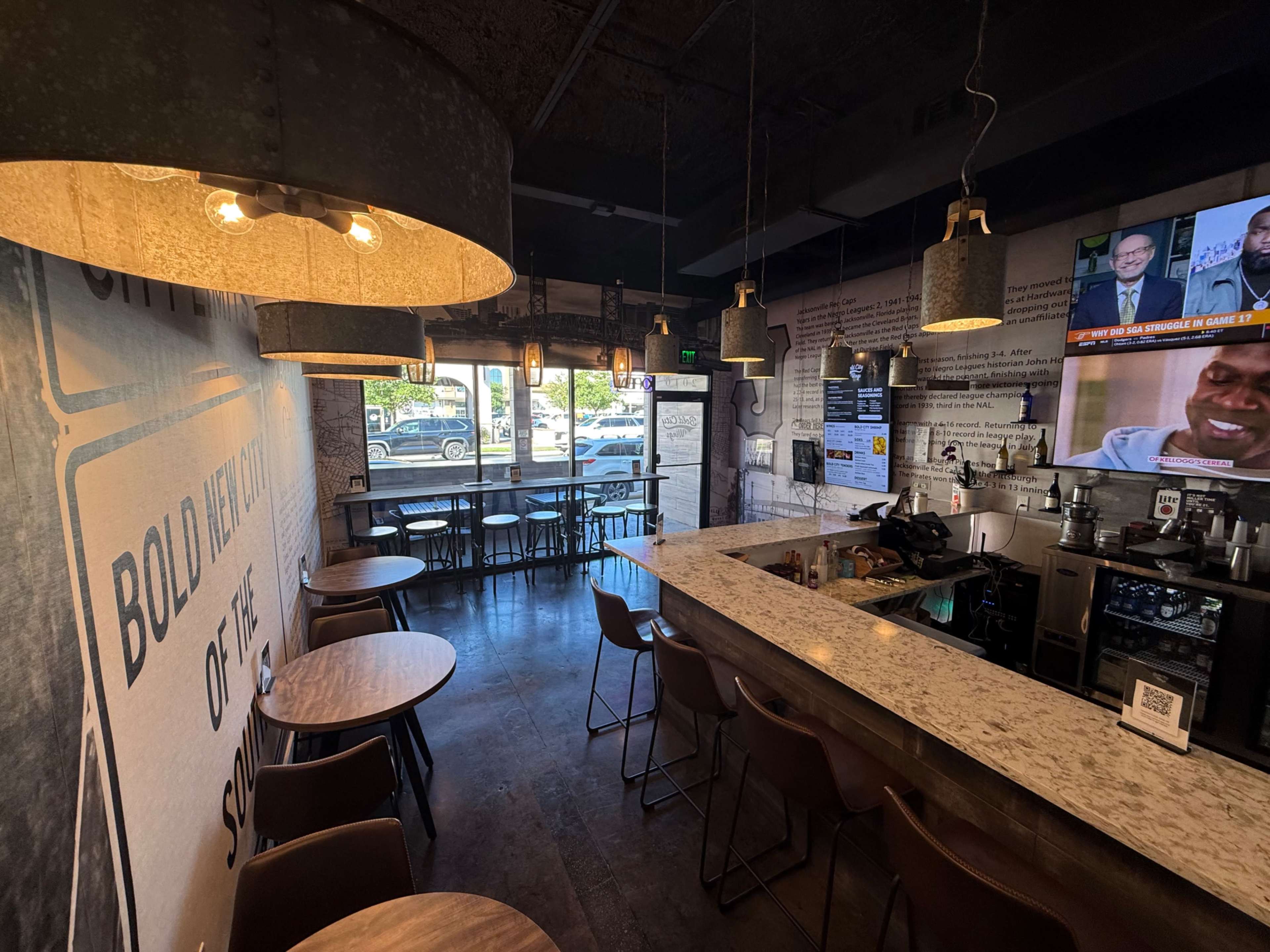 The image shows a cozy café interior with a bar area, wooden tables, and large pendant lights, along with a television displaying news in the background.