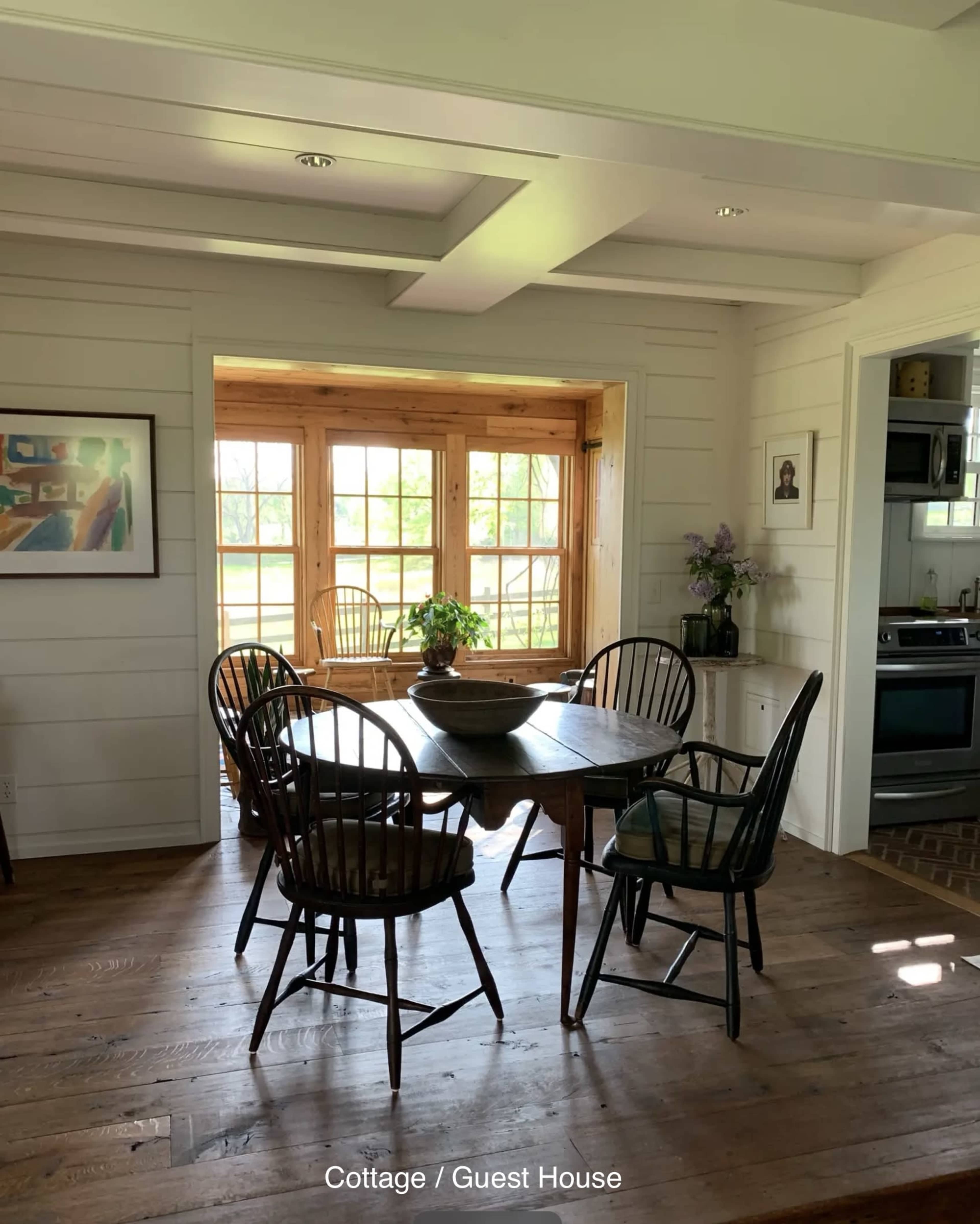 The image shows a cozy dining area in a cottage, featuring a round wooden table surrounded by four chairs, with a view of a sunlit room through large windows.