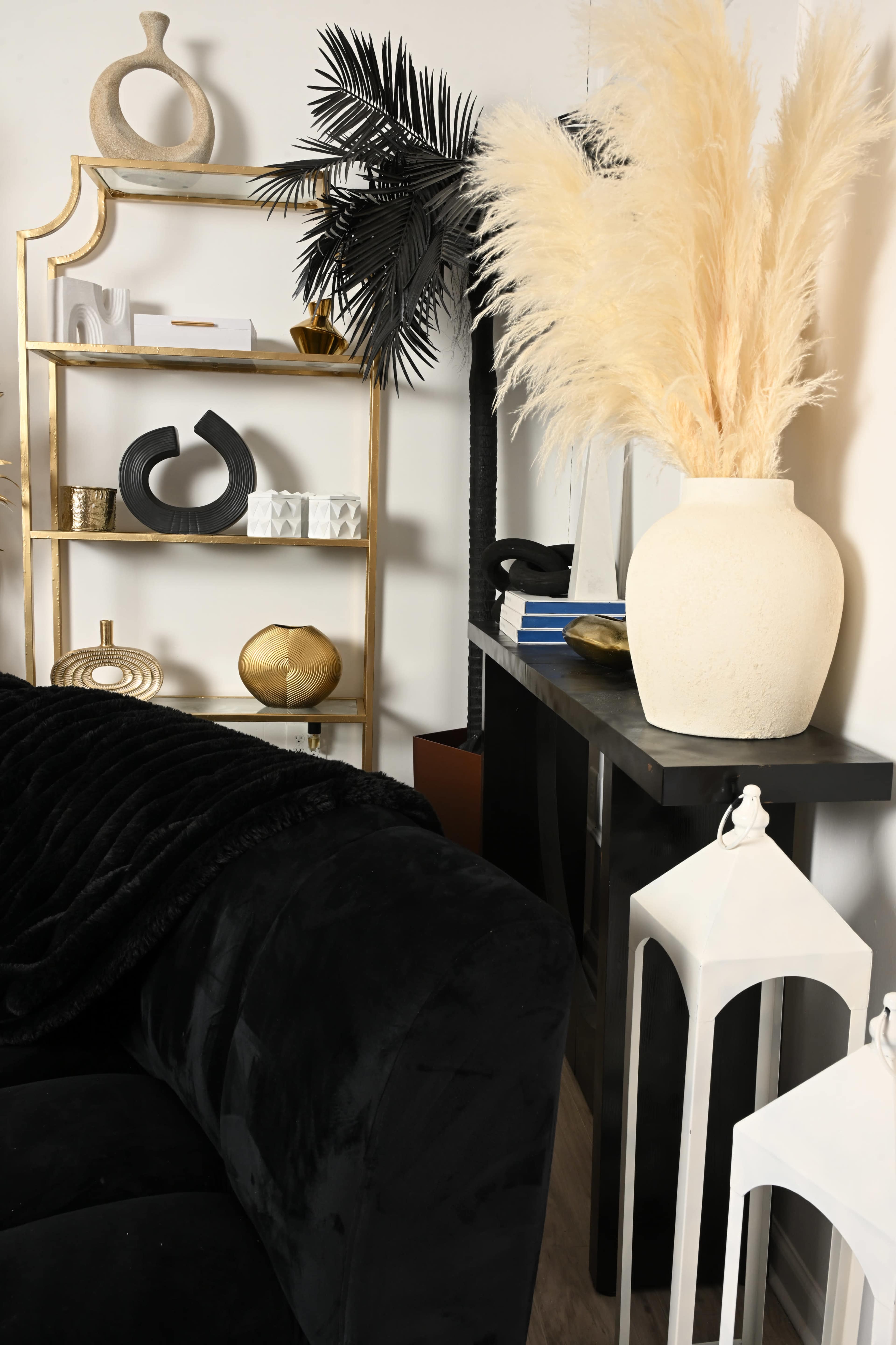 The image features a stylish interior scene with a black velvet sofa, a gold shelving unit displaying decorative items, and a large white vase filled with pampas grass beside a slender black console table.