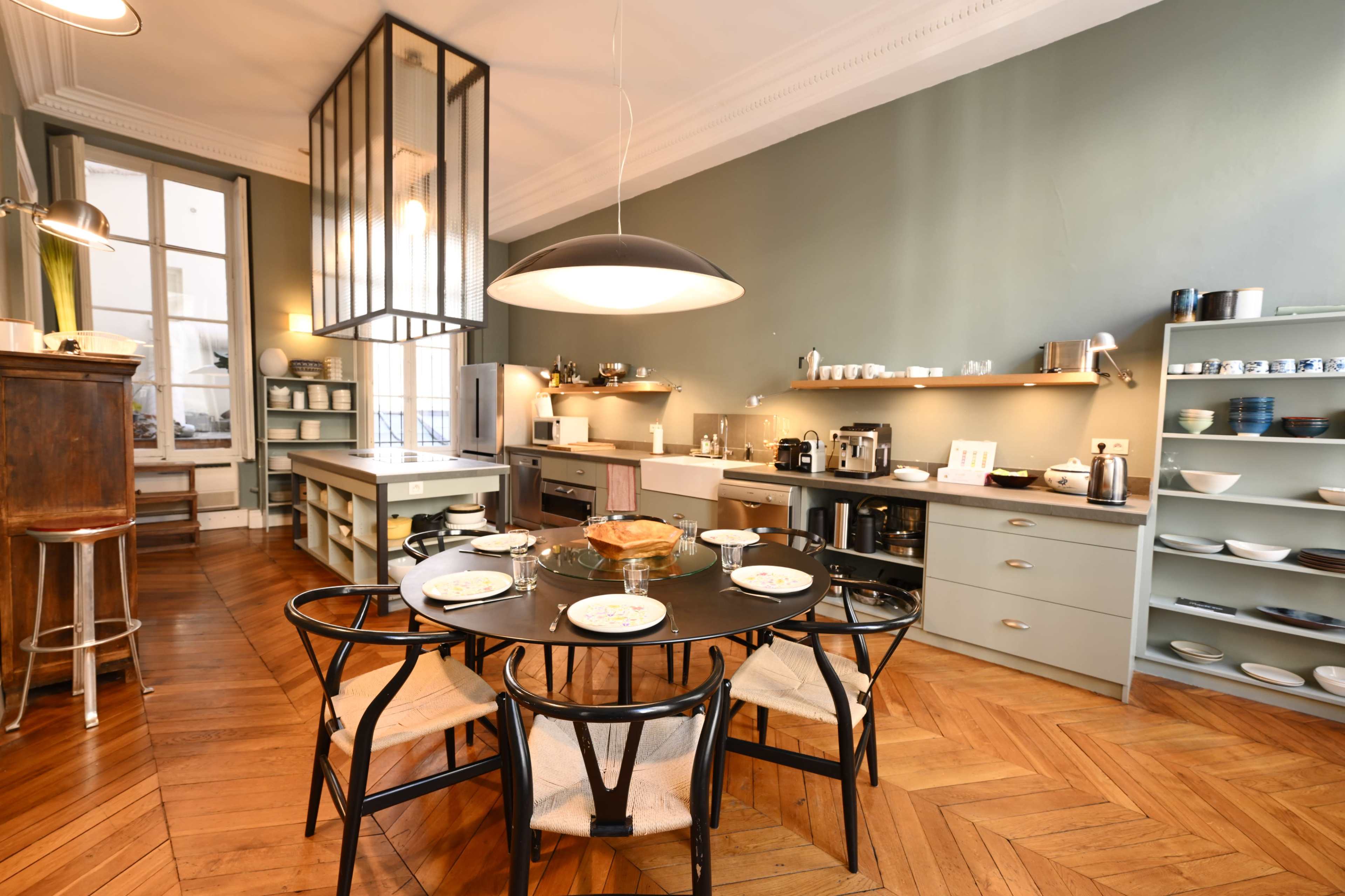 The image shows a modern kitchen and dining area featuring a round table set for a meal, with a well-organized kitchen layout and various kitchen appliances on display.