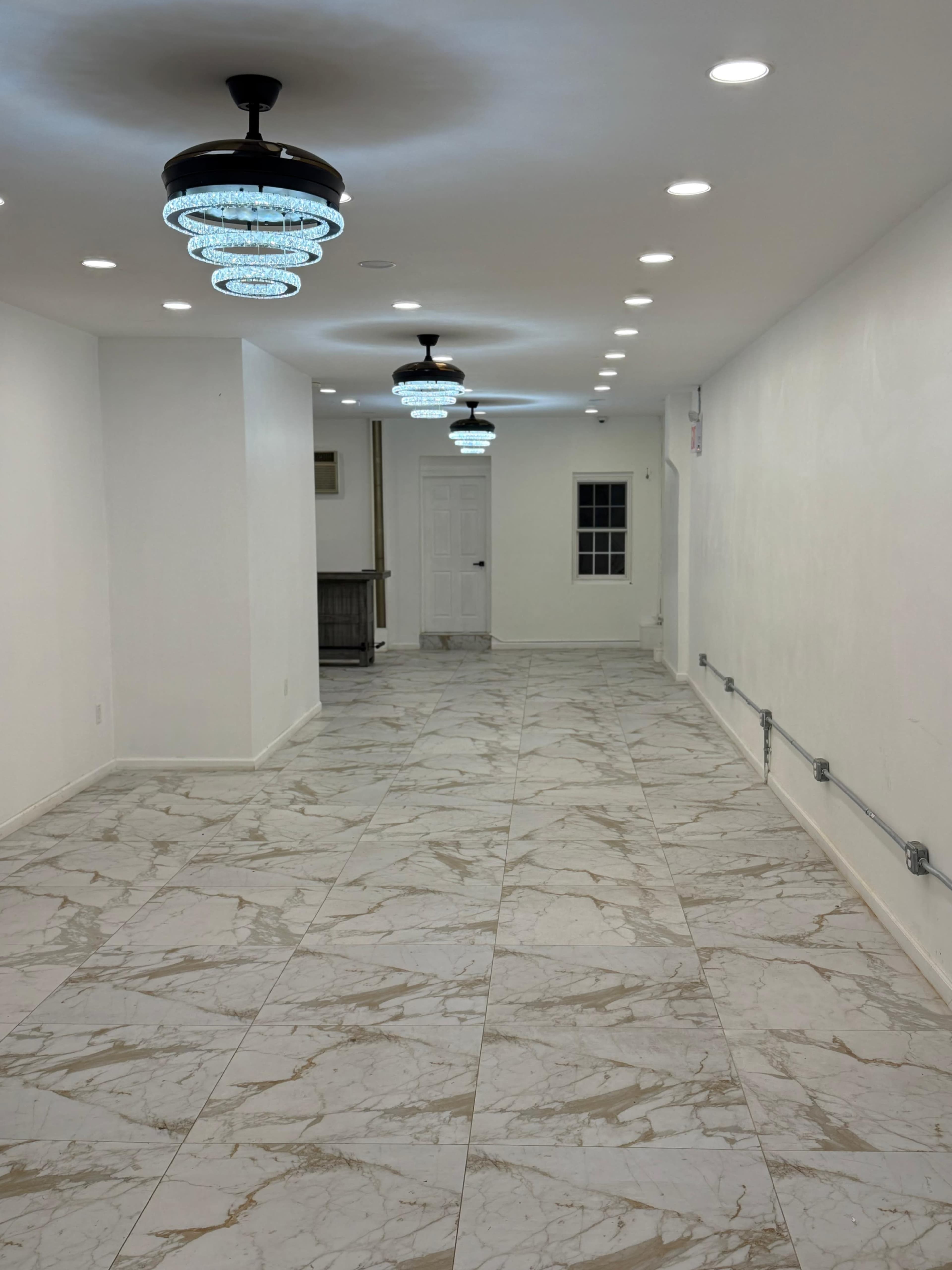 The image shows a long, narrow hallway with marble tile flooring and modern chandelier light fixtures illuminating the white walls.