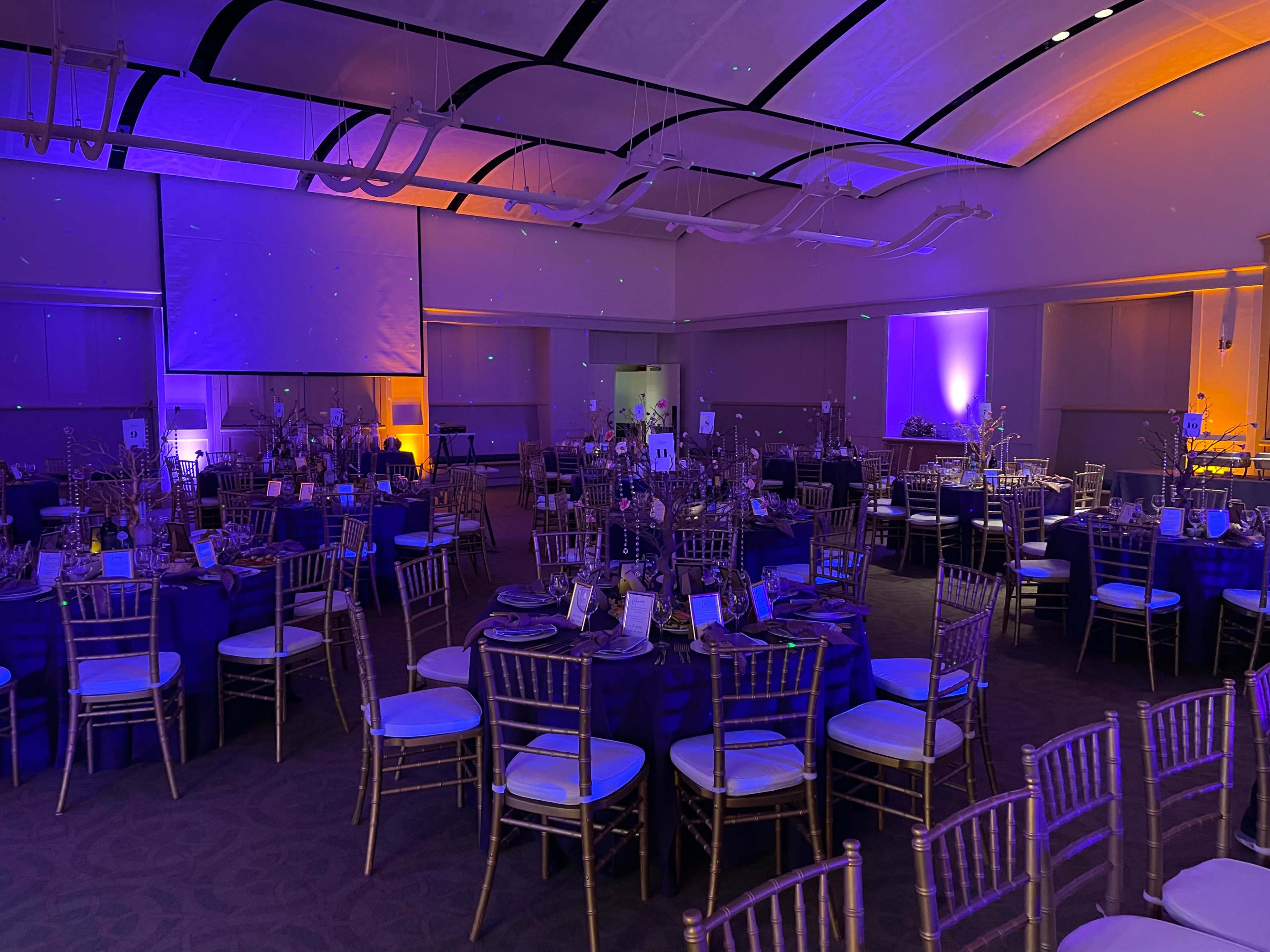 A banquet hall set up for an event, featuring round tables with blue tablecloths and gold chiavari chairs, illuminated by purple and orange lighting.