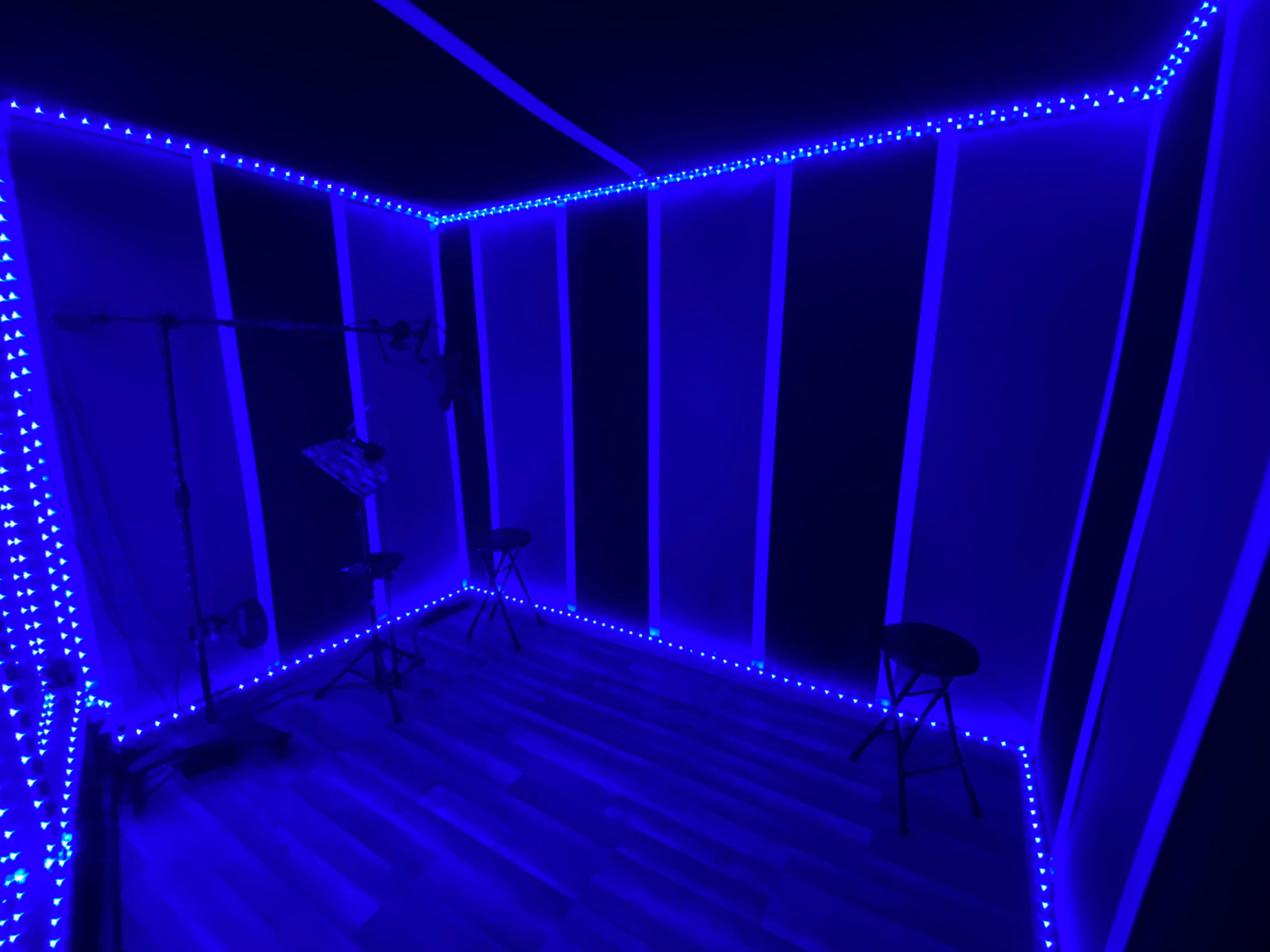 A sound recording studio with blue LED lights outlining the walls and two black stools positioned on a wooden floor.