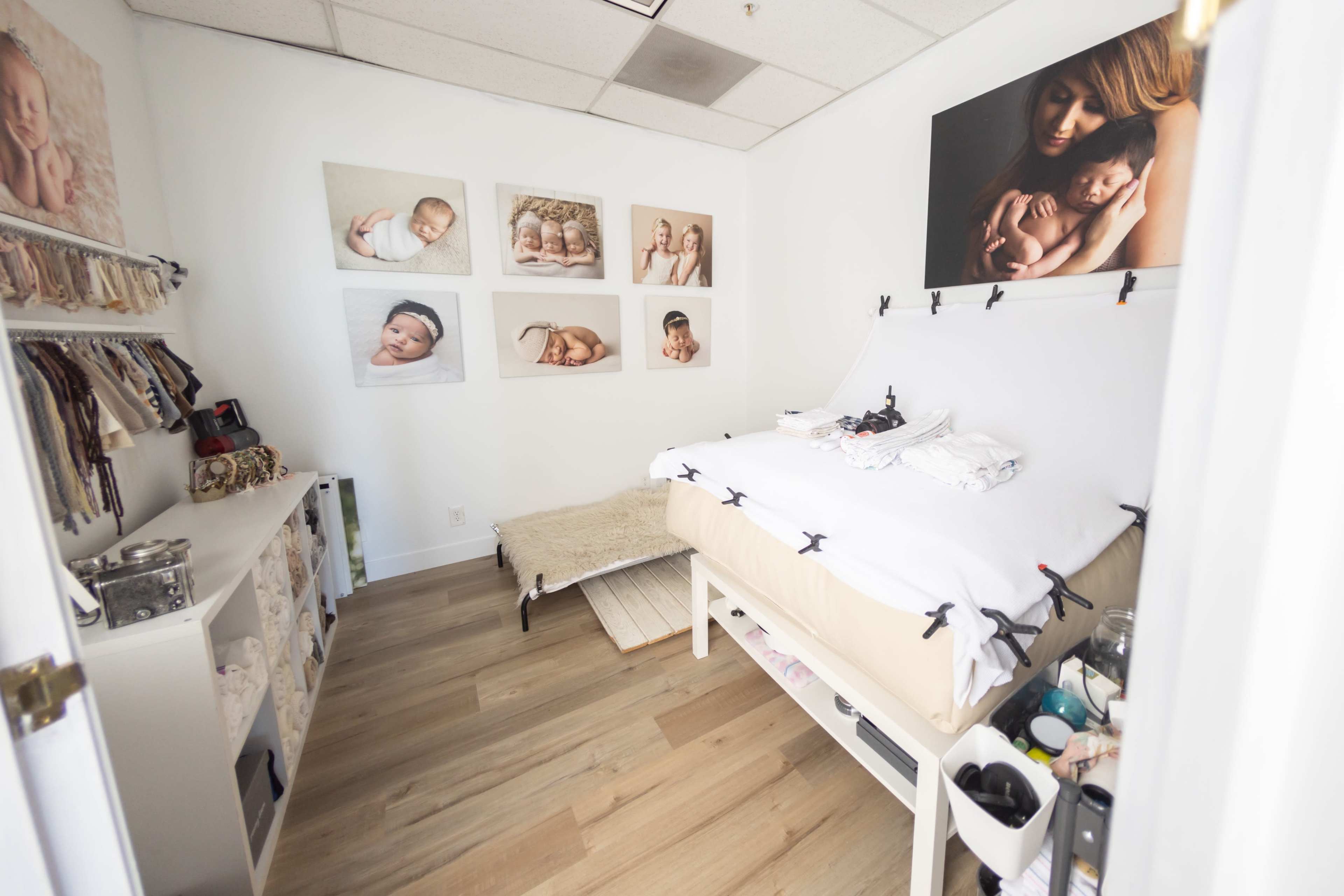 The image depicts a small studio or photography room featuring a bed draped with white sheets, surrounded by multiple framed photos of infants on the walls.