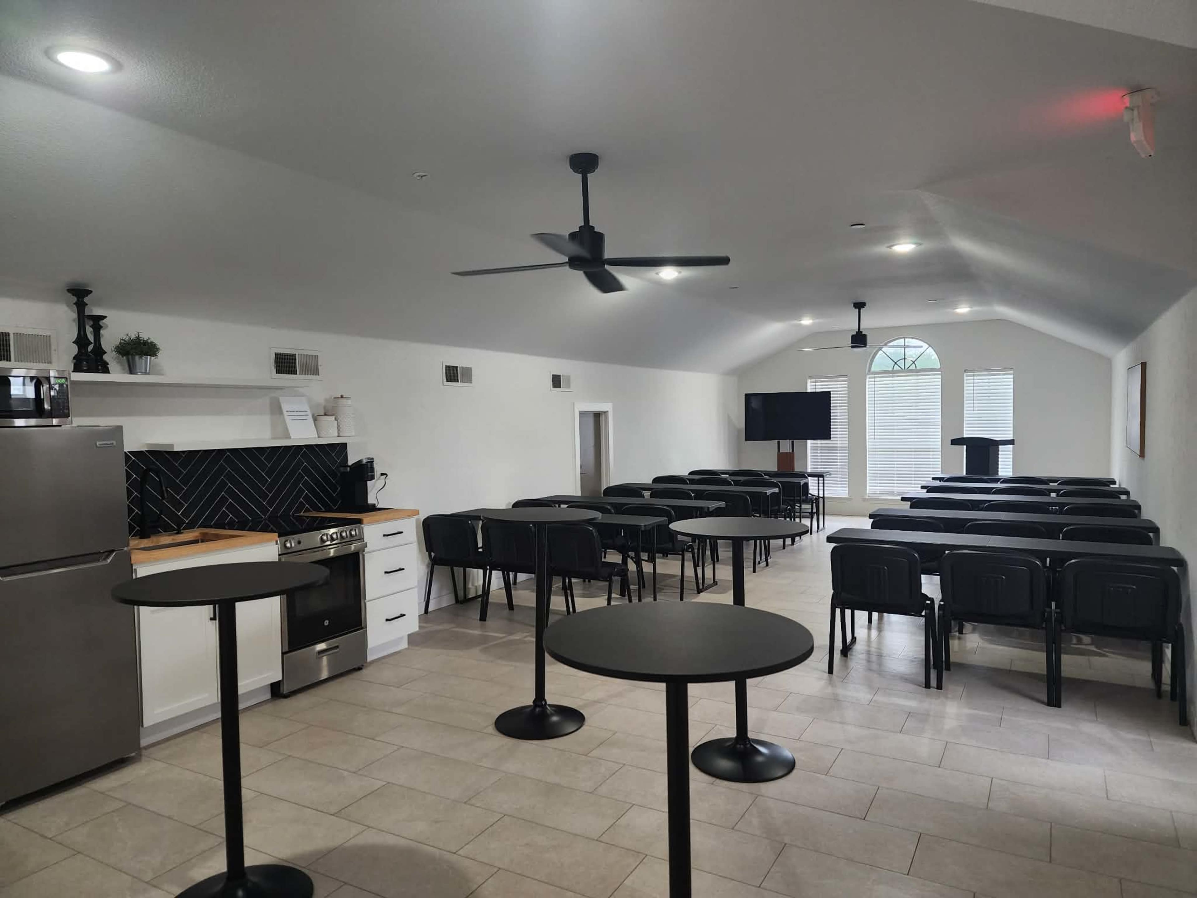 The image shows a spacious meeting room with several rows of black chairs, round tables, a kitchen area, and large windows allowing natural light to enter.