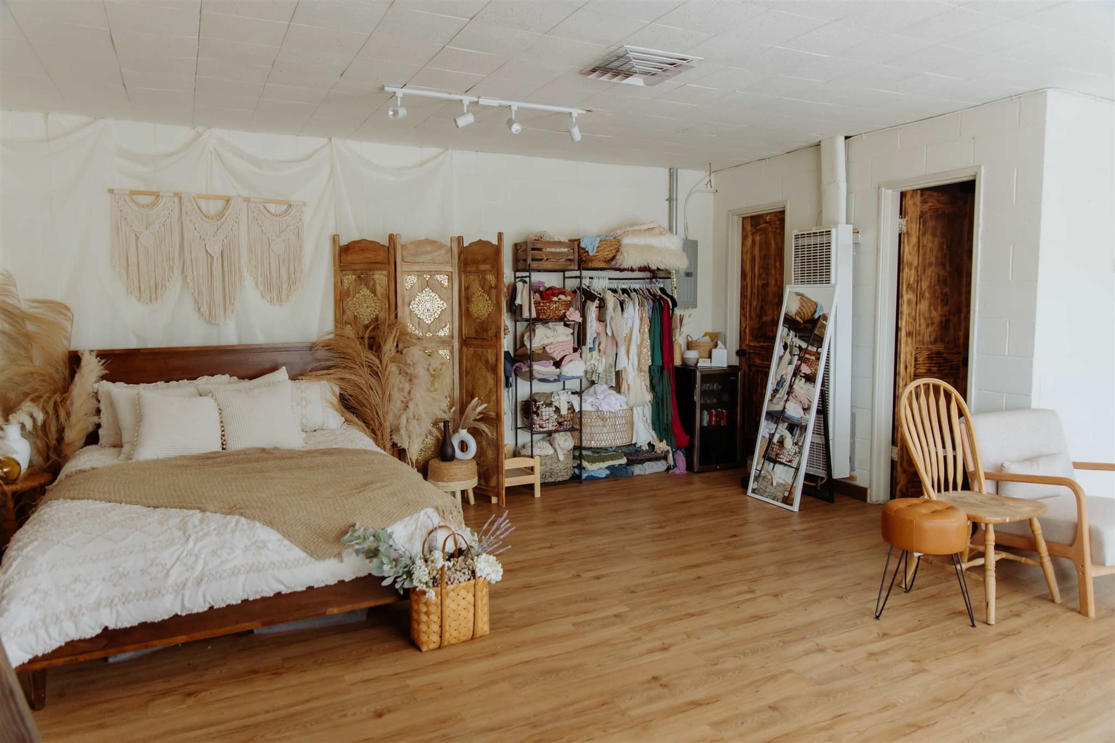 The image shows a cozy, well-decorated bedroom with a wooden bed, a clothing rack, a large mirror, and a chair arranged in a spacious area with wooden flooring.