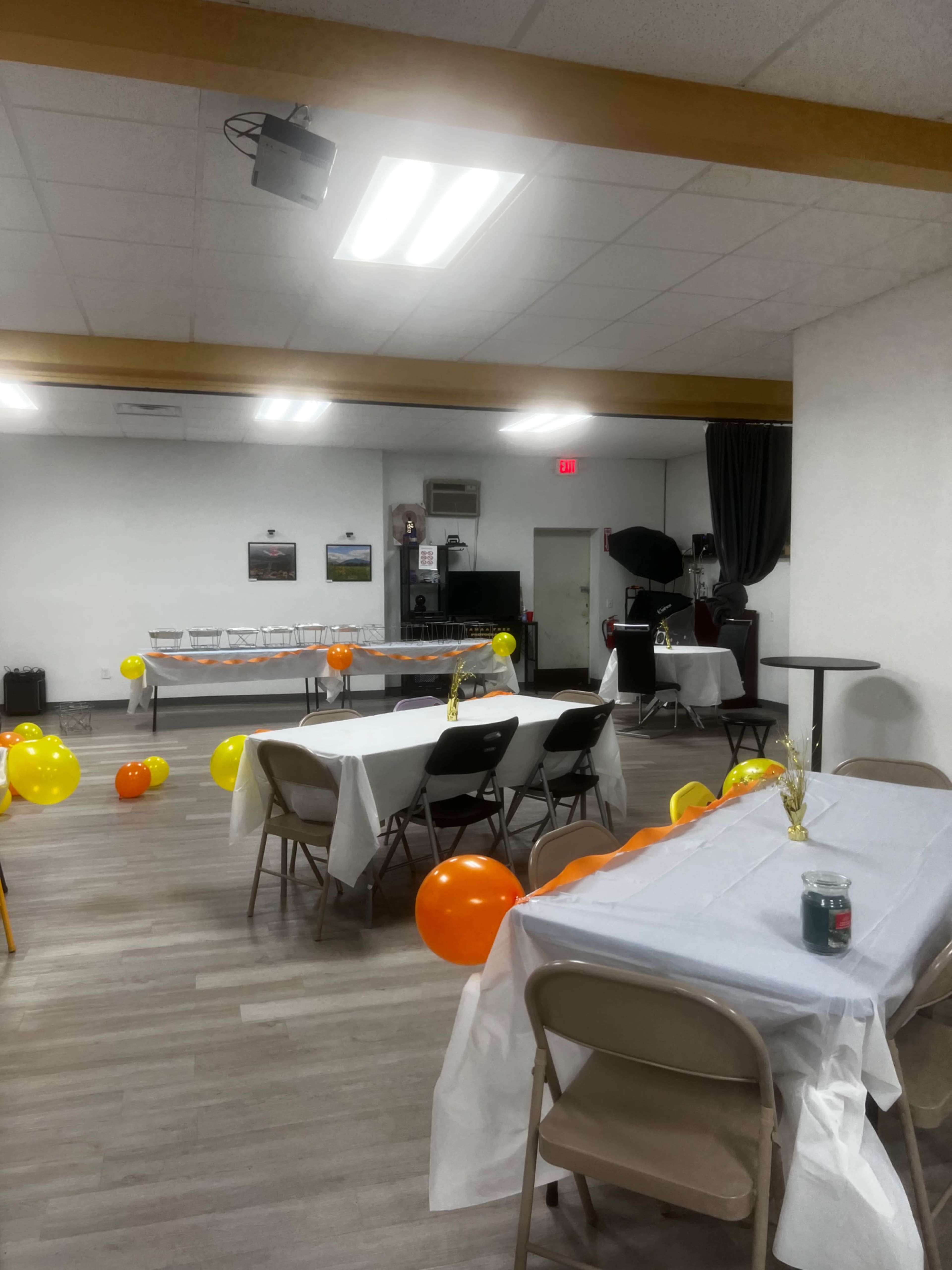 The image shows a banquet hall decorated with orange and yellow balloons, featuring several tables covered with white tablecloths arranged for an event.