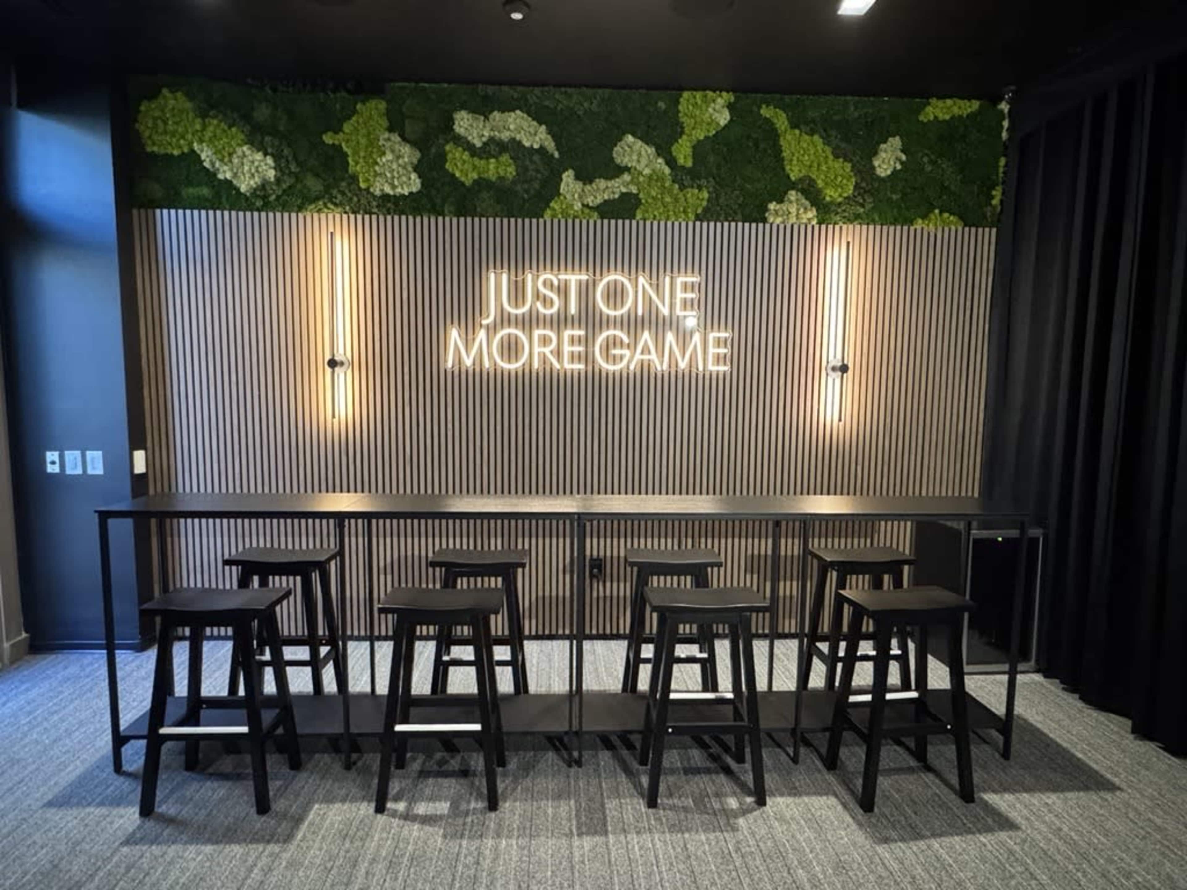 The image shows a modern game room interior featuring a bar-like table with several black stools and a neon sign that reads "JUST ONE MORE GAME" on a textured wall with greenery above.