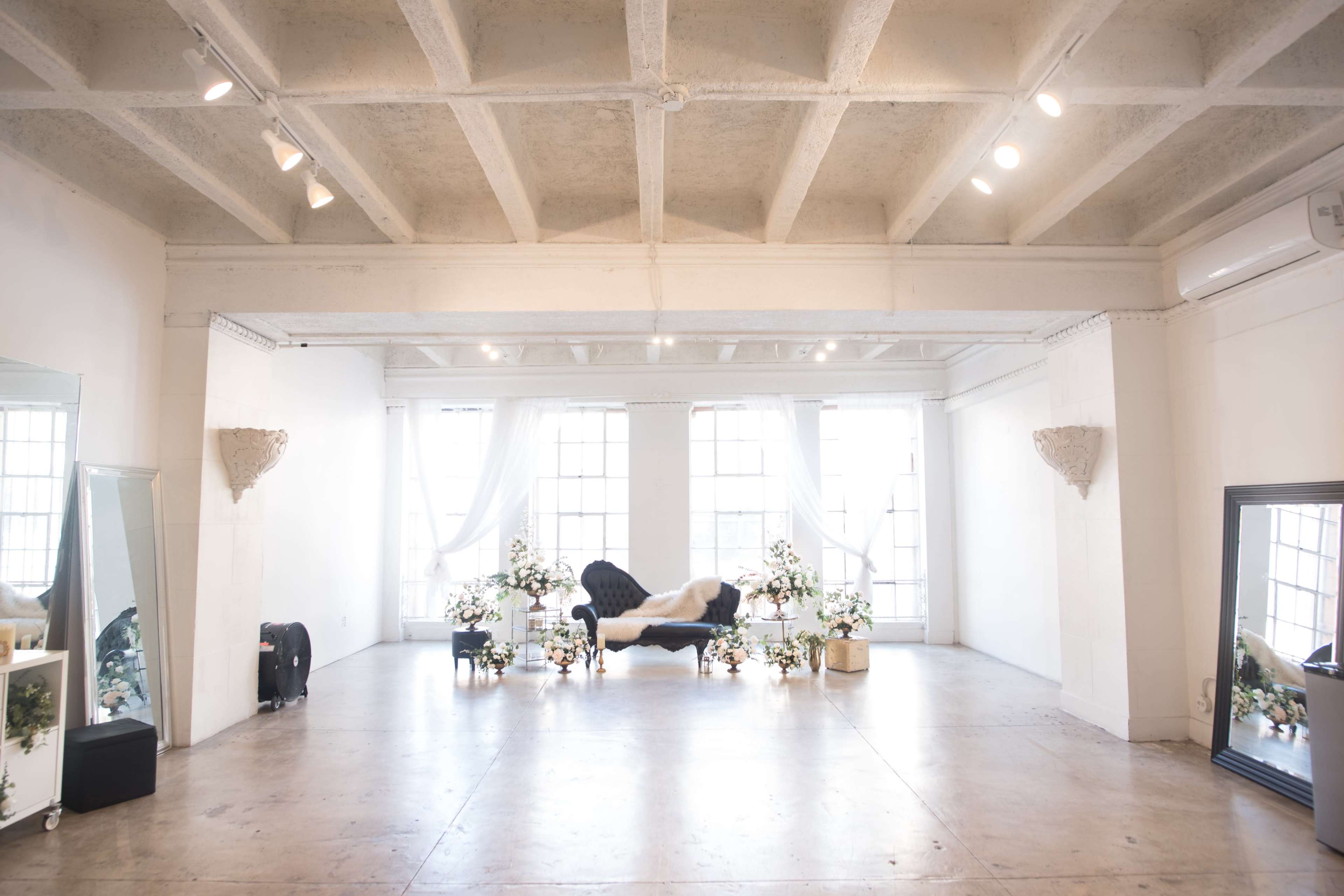 A spacious room features a dark blue couch surrounded by potted plants and large windows, with tall mirrors positioned on either side.