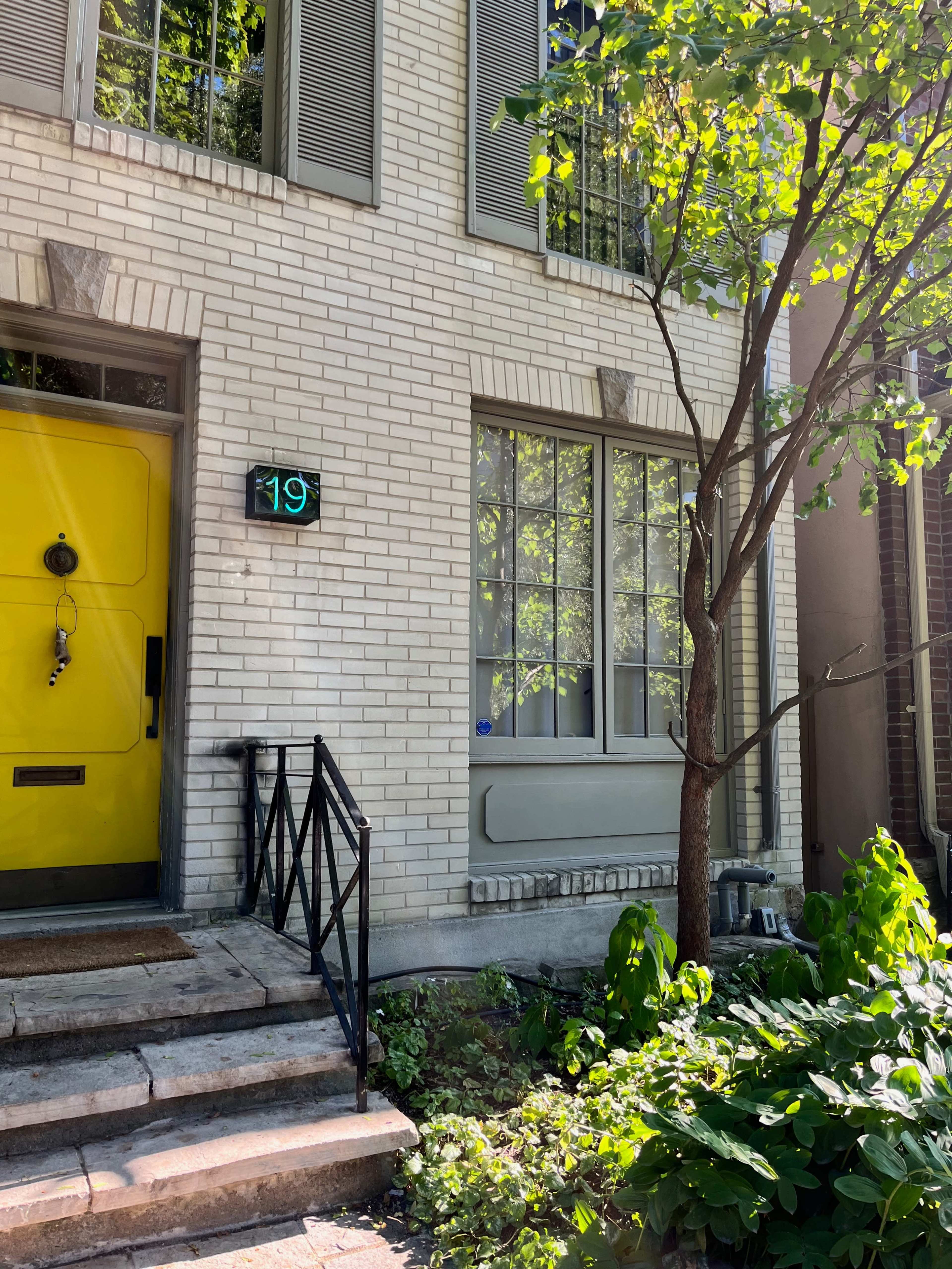 A light yellow front door with the number 19 displayed next to it, framed by white brick walls and a small garden with green plants.