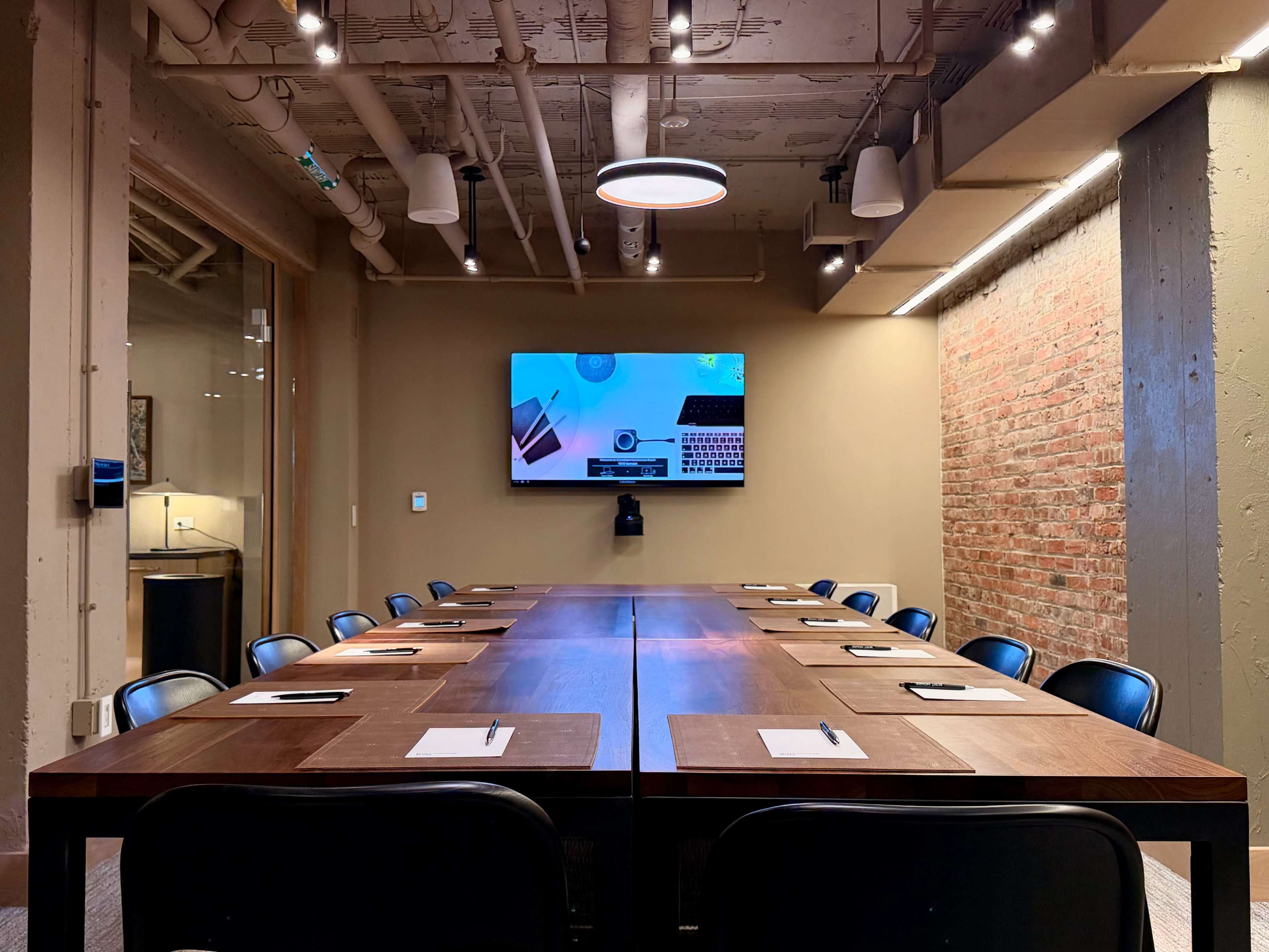 A rectangular conference table with ten chairs is set up in a room featuring a large screen mounted on the wall, exposed brick, and overhead lighting.
