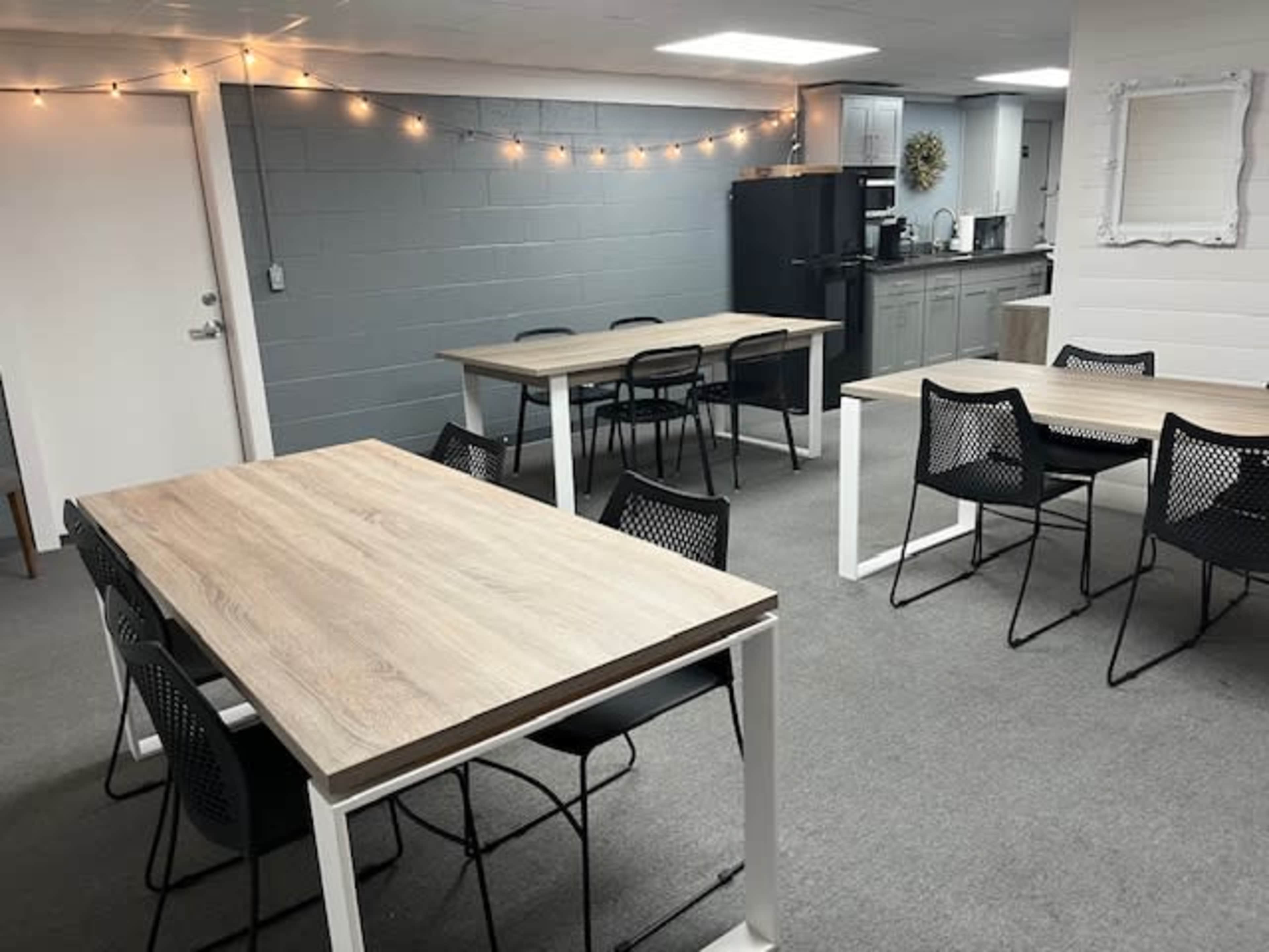A room with several wooden tables and chairs arranged, along with a kitchenette area featuring a black refrigerator and light fixtures along the wall.