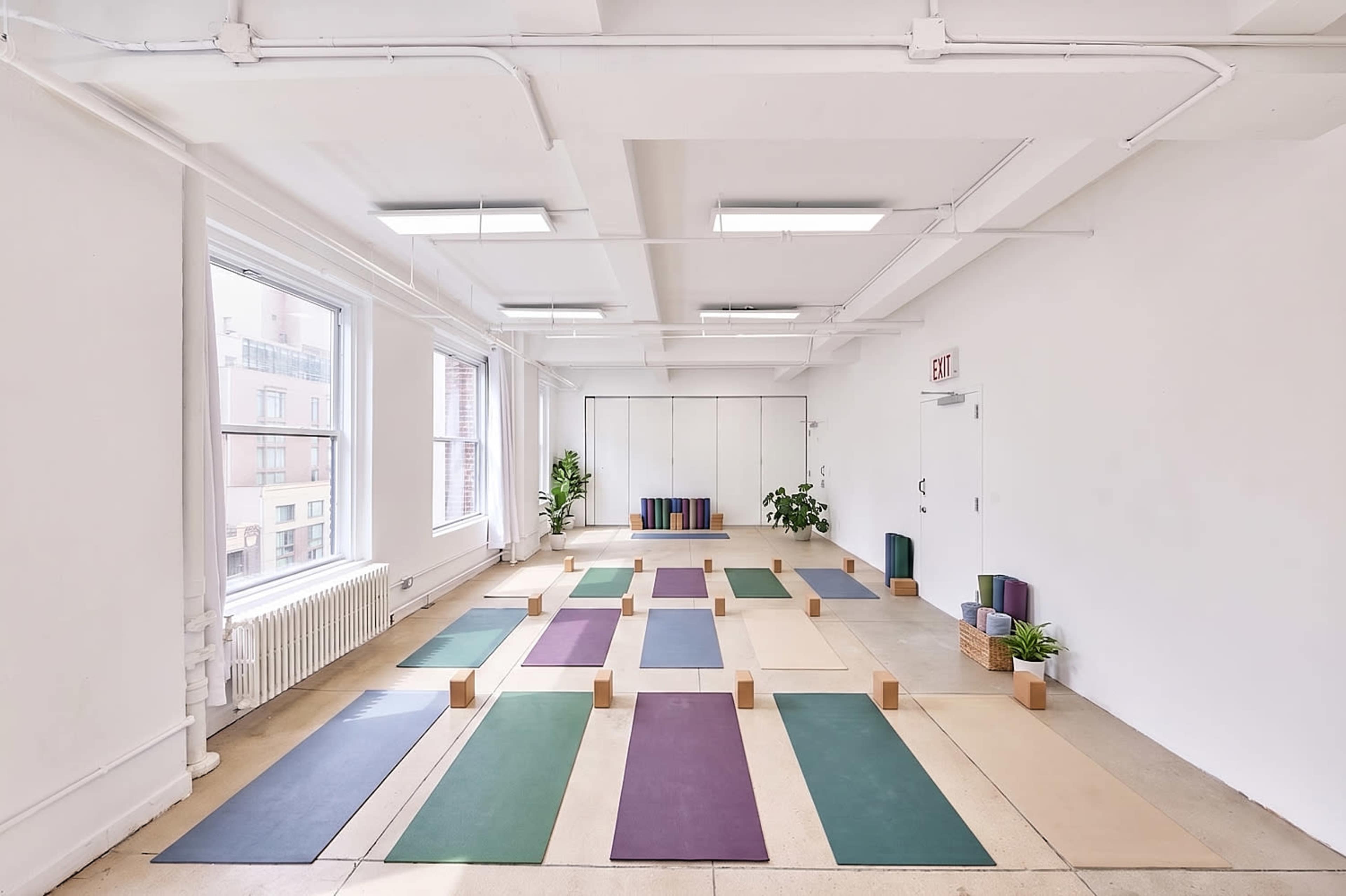 The image shows a spacious yoga studio with yoga mats arranged in a grid pattern, wooden blocks, and potted plants against plain white walls.