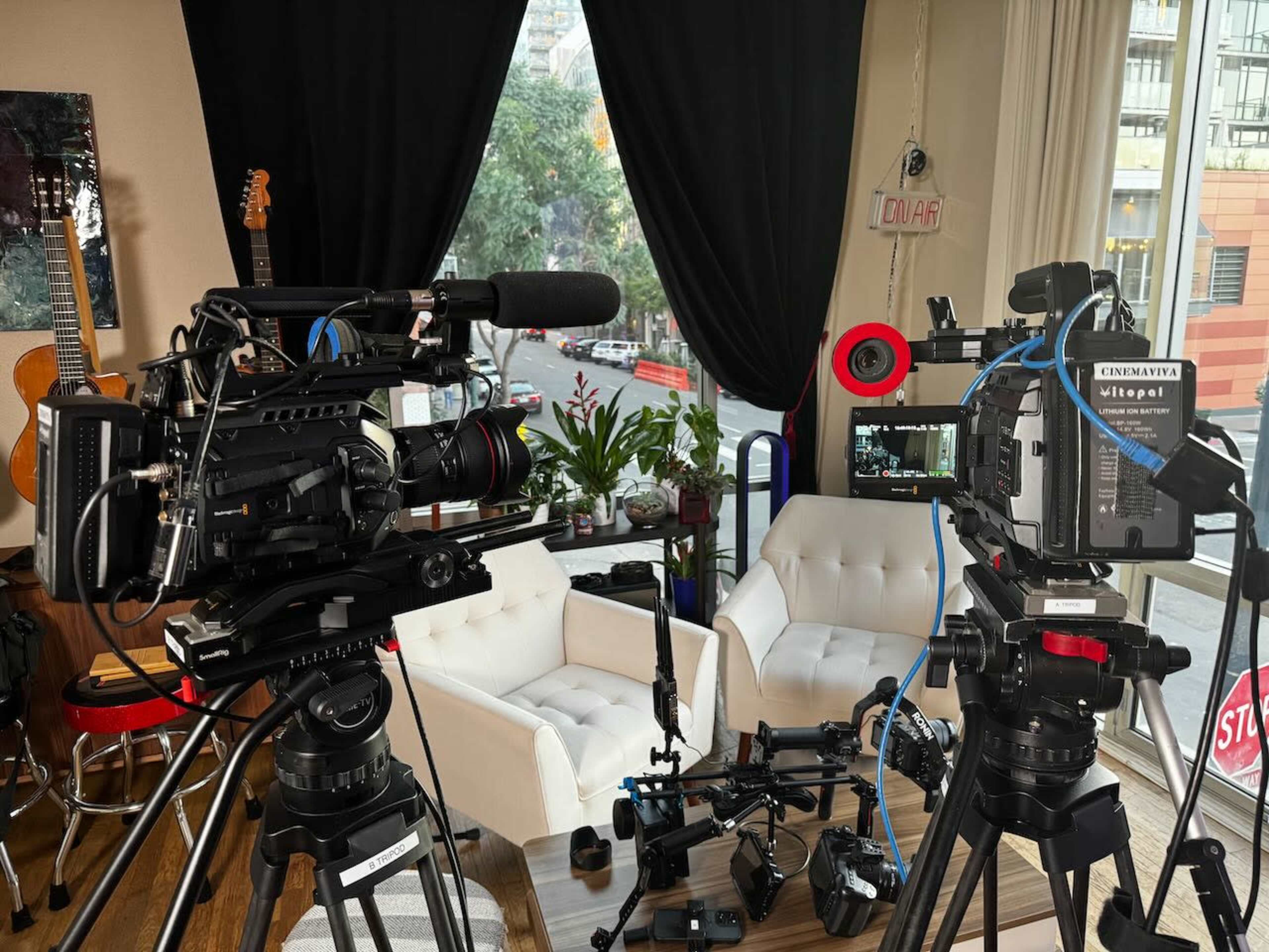 The image shows two video cameras on tripods facing a pair of white armchairs in a well-lit interior space with a window and some greenery outside.