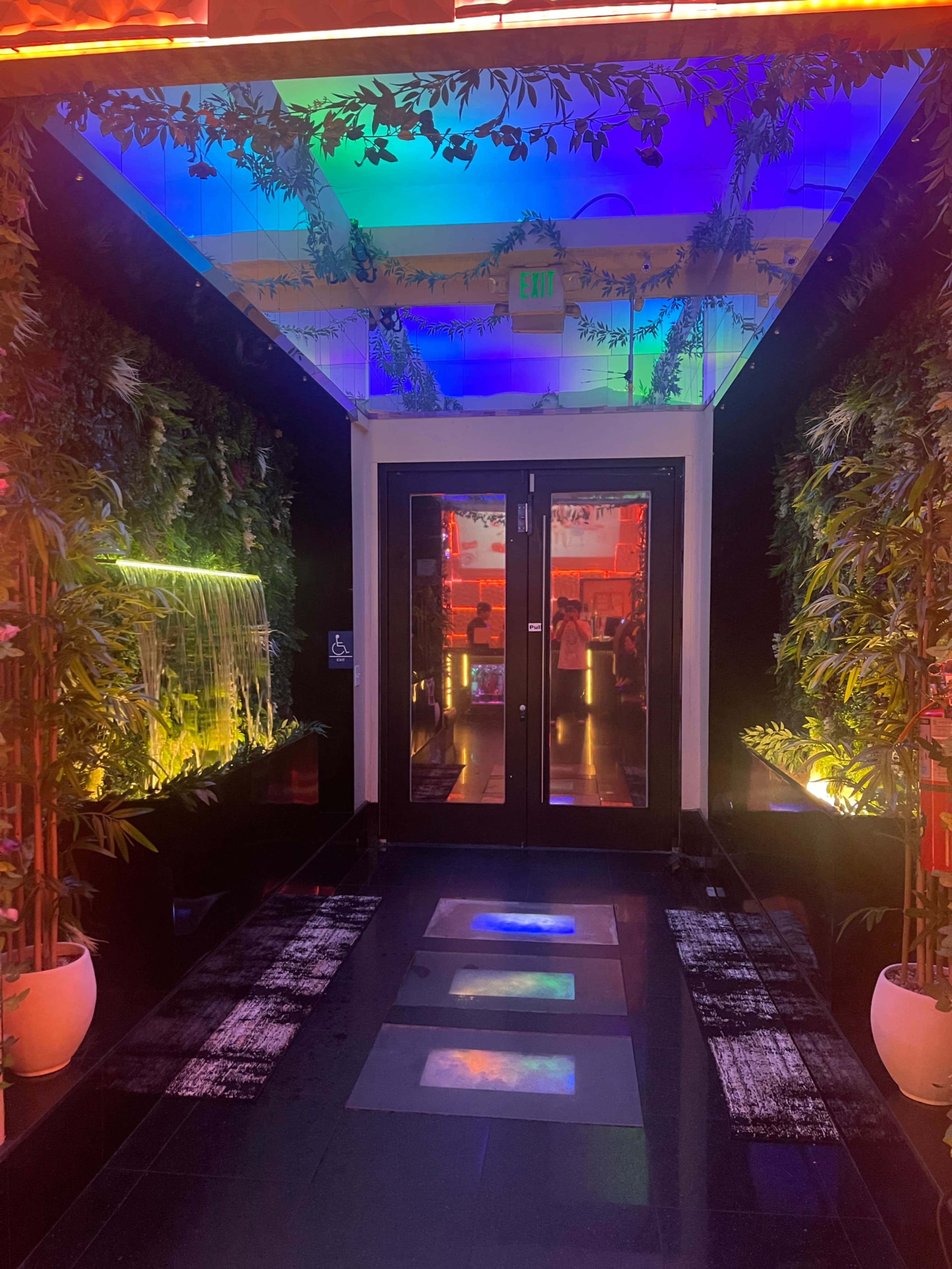 A vibrant hallway features colorful lighting, greenery, and a small waterfall, leading to double glass doors.