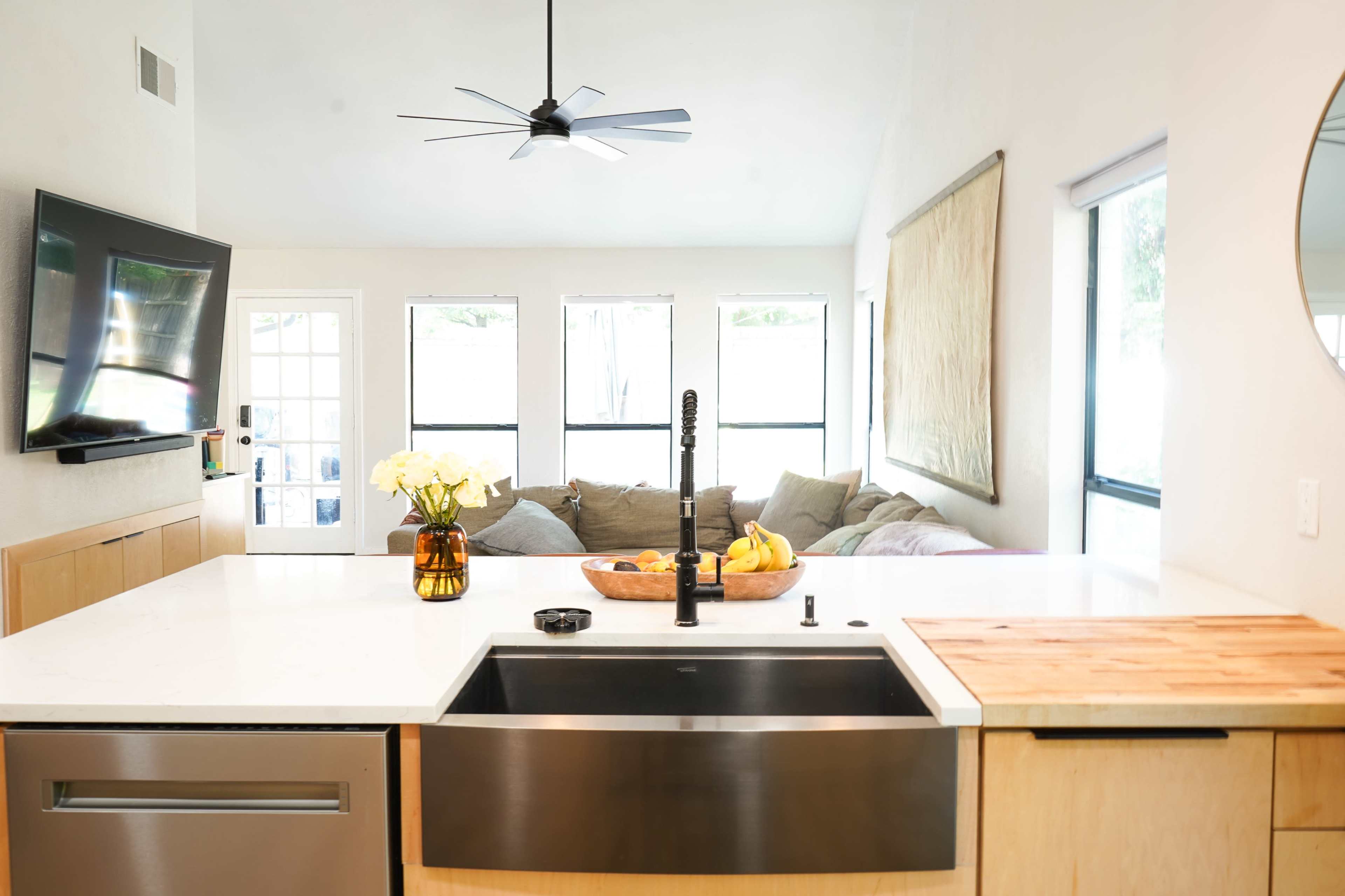 A modern kitchen features a stainless steel sink, a wooden cutting board, and a countertop adorned with a vase of flowers and a bowl of bananas, with a living area visible in the background.