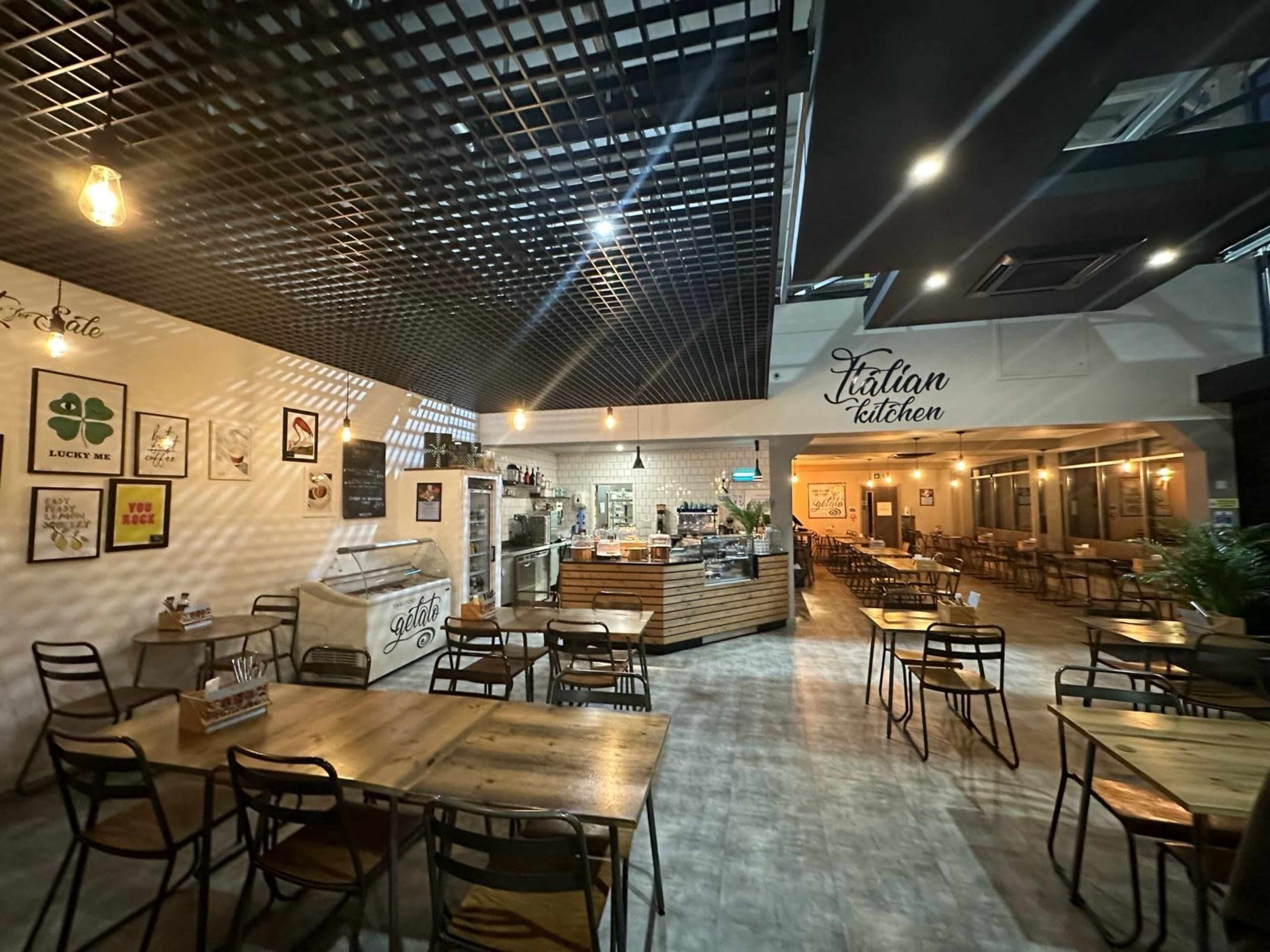 A spacious Italian restaurant interior featuring wooden tables, metal chairs, and a counter with a coffee machine and displayed food items.