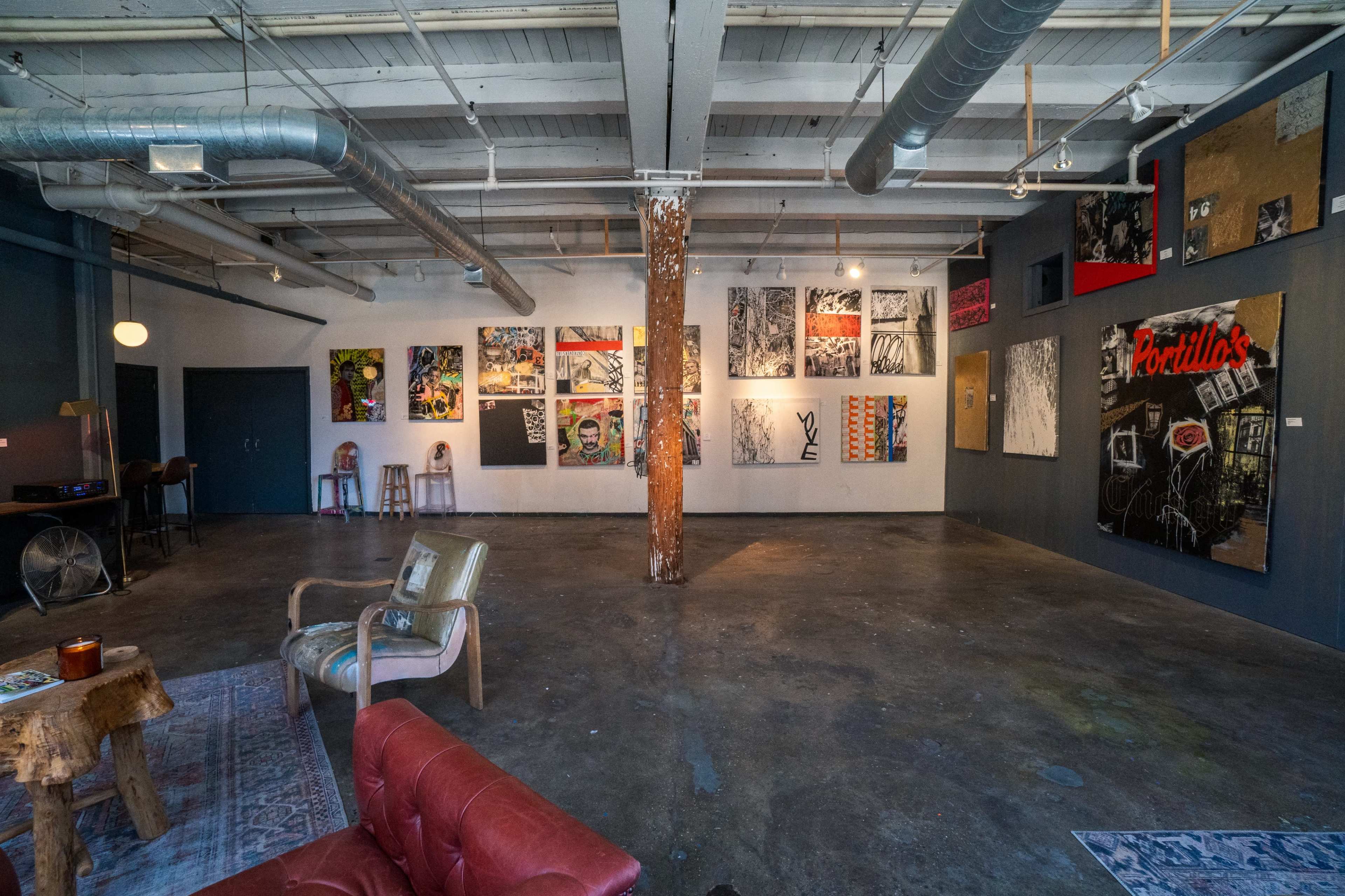 A spacious art gallery with concrete flooring, featuring a variety of colorful artworks displayed on the walls, along with a seating area.