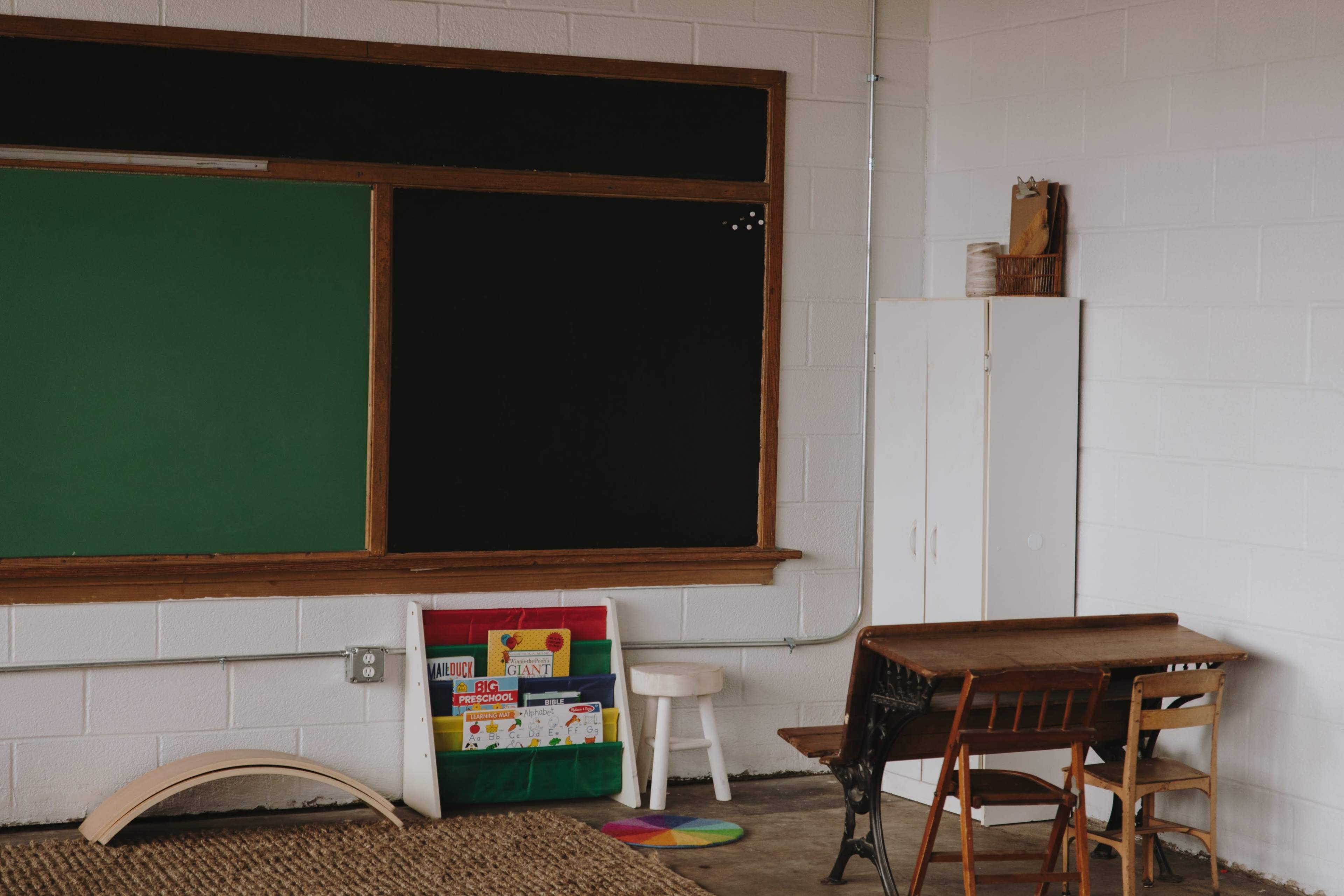 The image shows a classroom with a green chalkboard, a blackboard, a wooden desk and chair, a white cabinet, and colorful books on a shelf nearby.