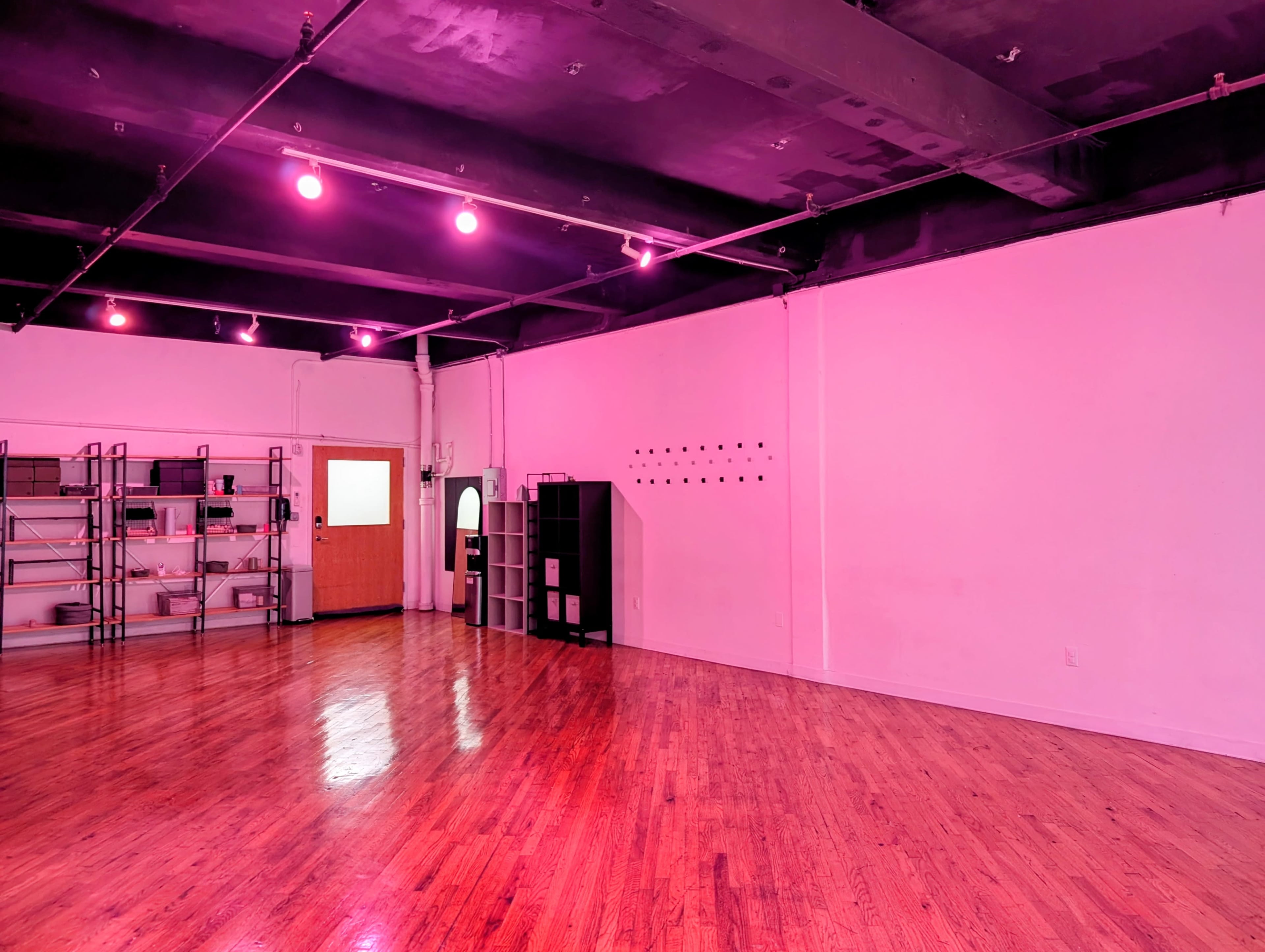 The image shows a spacious room with wooden floors, pink lighting, and minimal furniture, including shelves and a black storage unit against the wall.