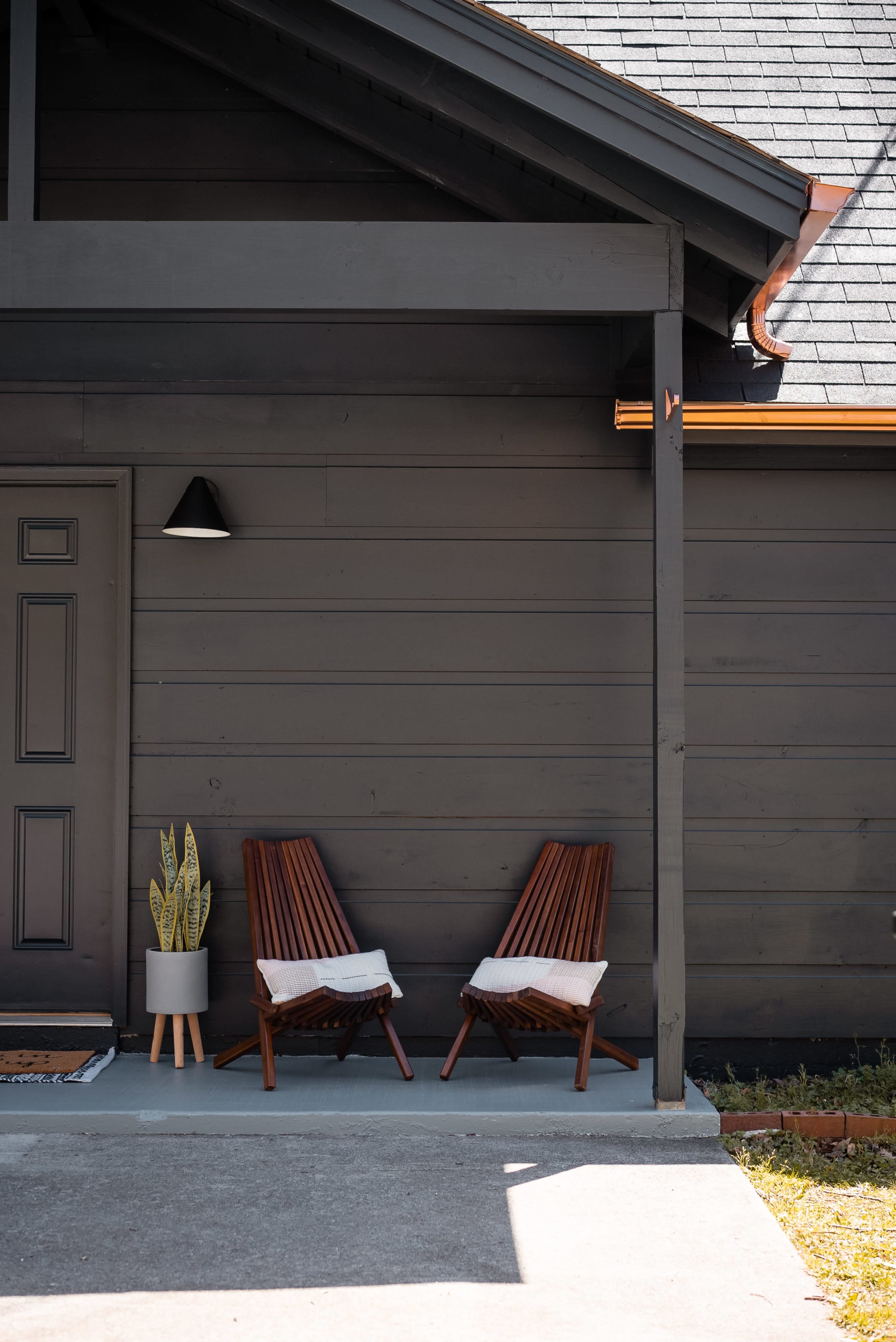 Two wooden lounge chairs with cushions are positioned on a concrete patio next to a potted plant against a dark-colored house exterior.