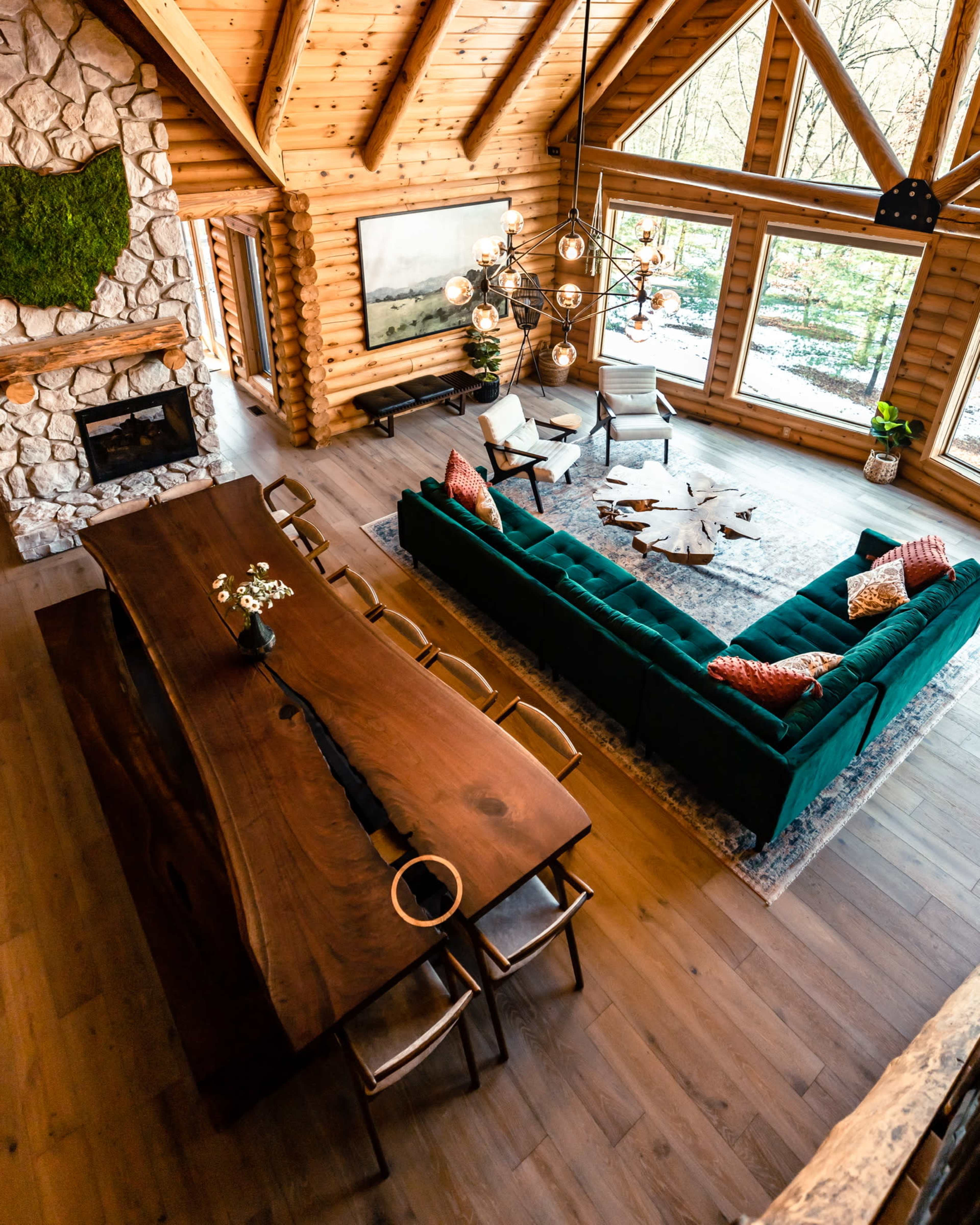 A spacious log cabin interior featuring a large wooden dining table, a green sofa arrangement, and a stone fireplace, with large windows providing views of the surrounding nature.