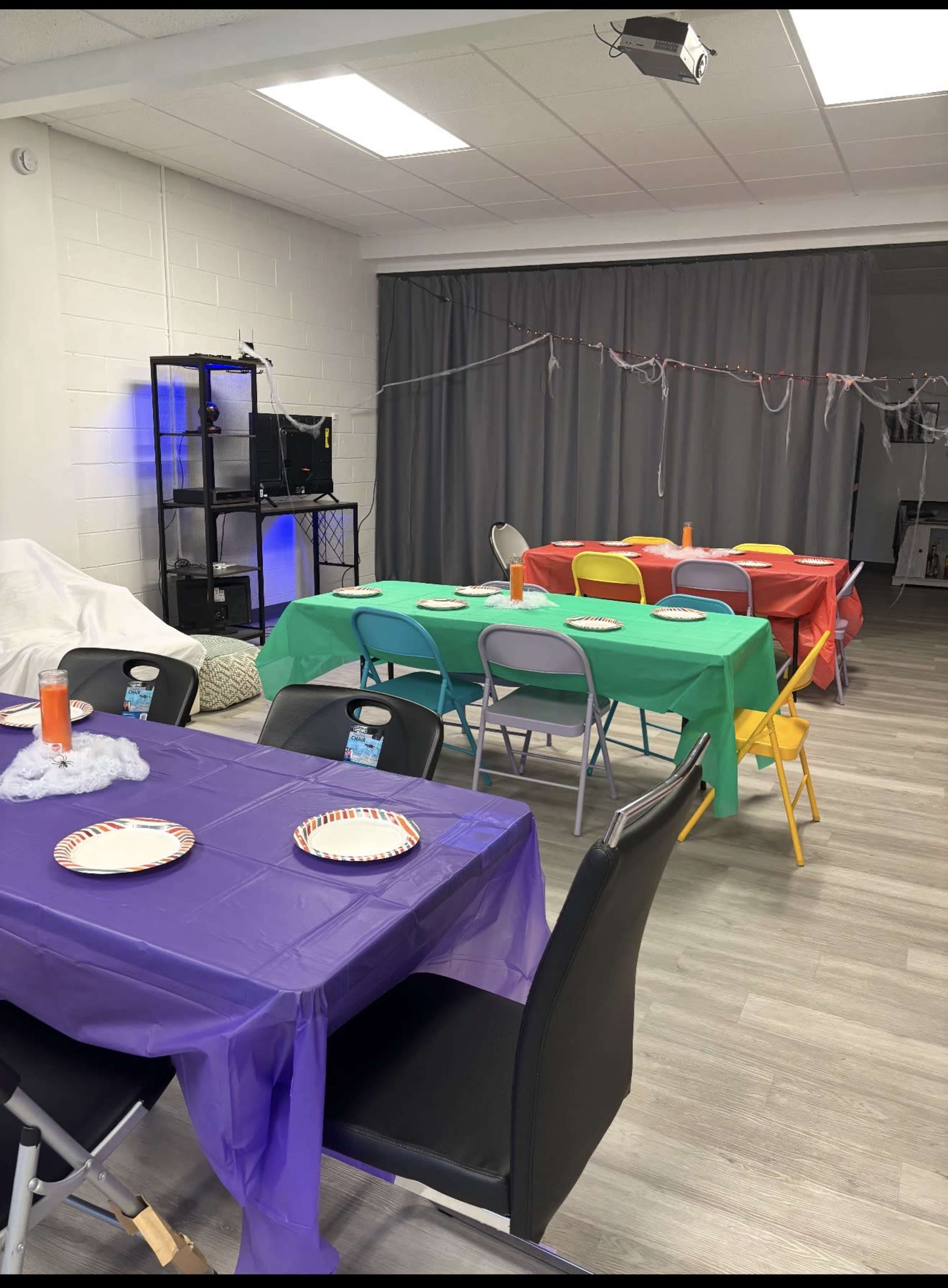 The image shows a room set up for a gathering, featuring several tables covered with colorful tablecloths, each with plates and drinks, and a gray curtain in the background.