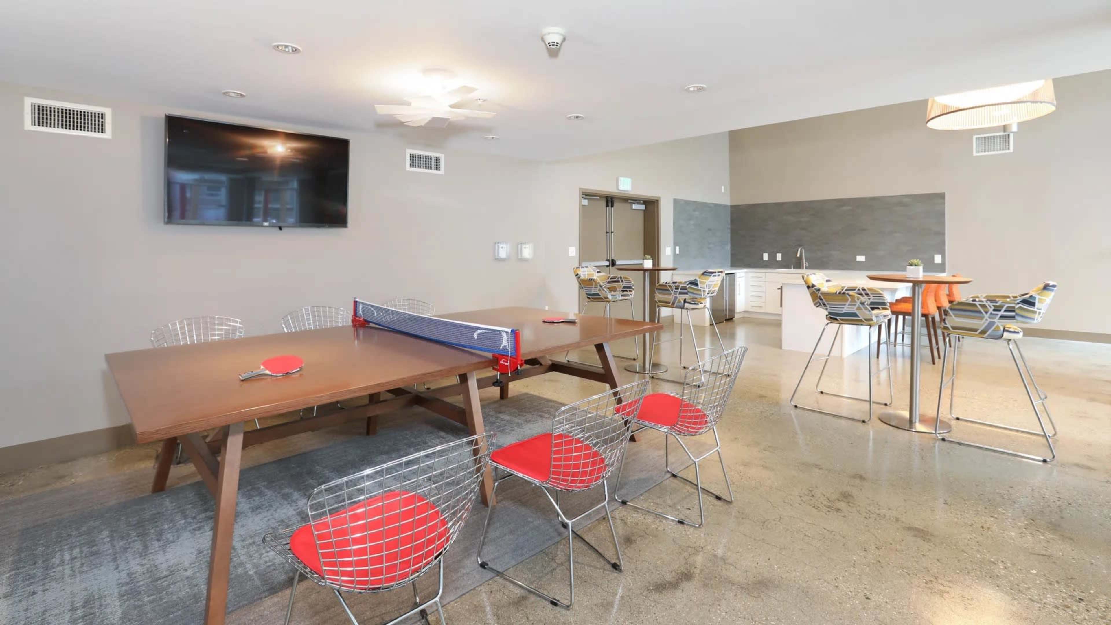 The image shows a modern communal lounge area with a table, chairs, a television, and a food preparation space.