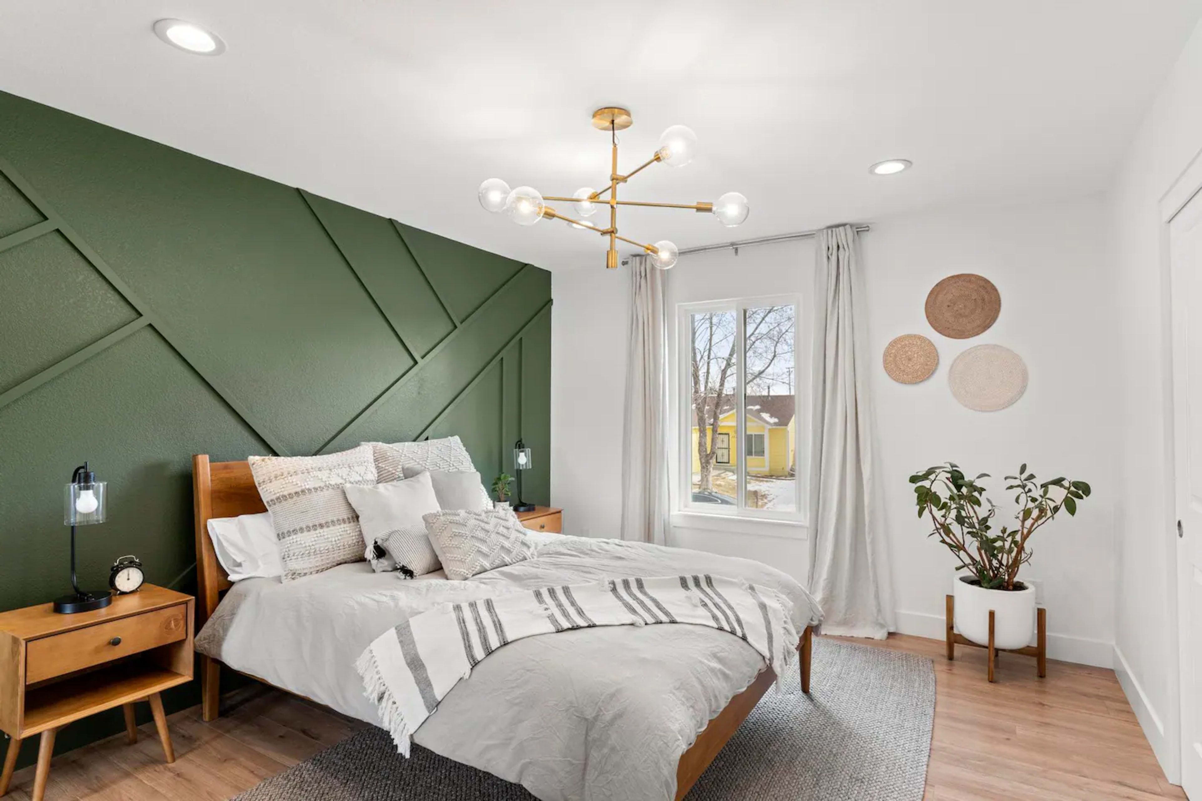 A modern bedroom with a green accent wall, a wooden bed with gray bedding, and a window overlooking an outdoor area.