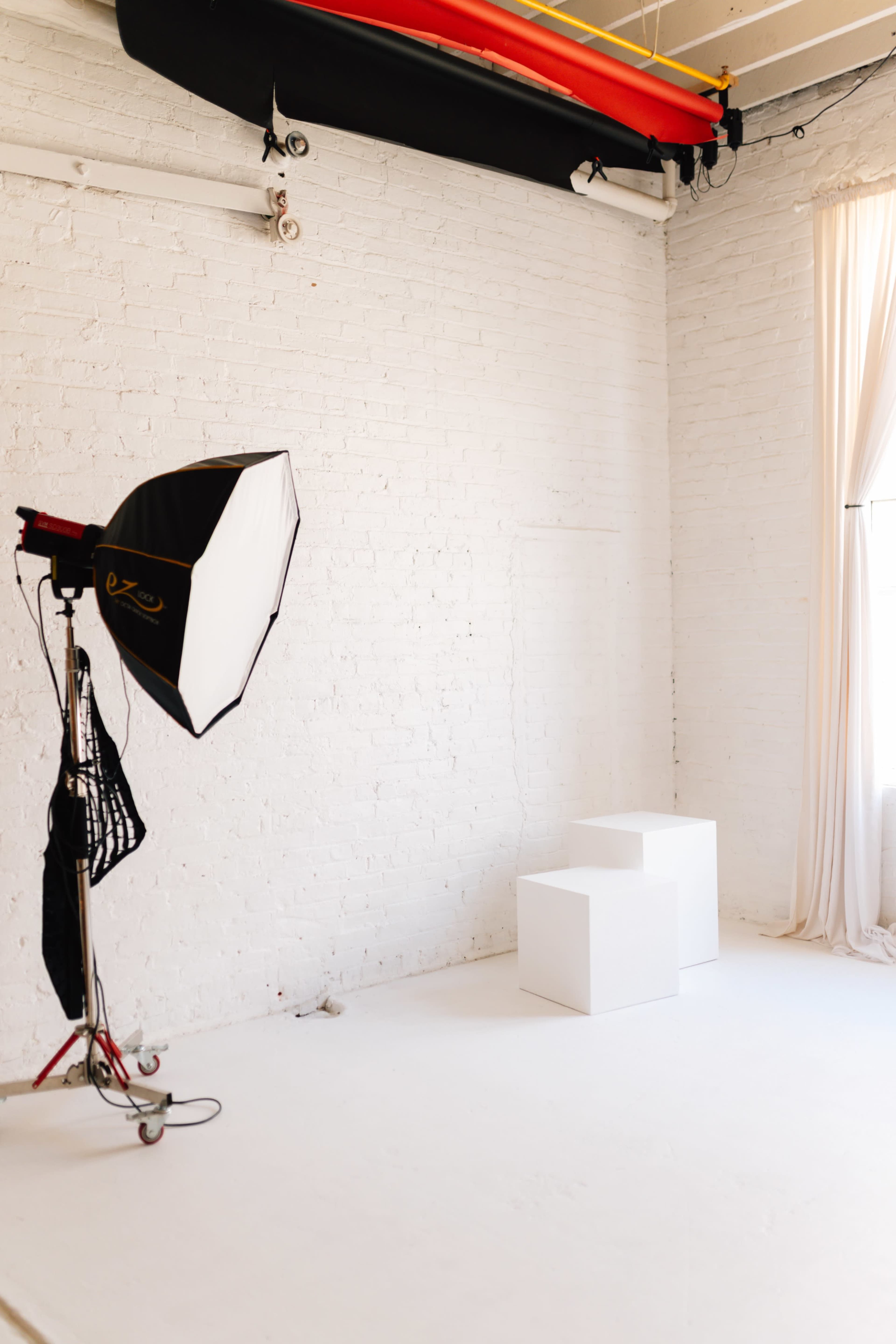 The image shows a minimalistic photography studio with two white blocks and a large softbox light set on a stand against a white brick wall.