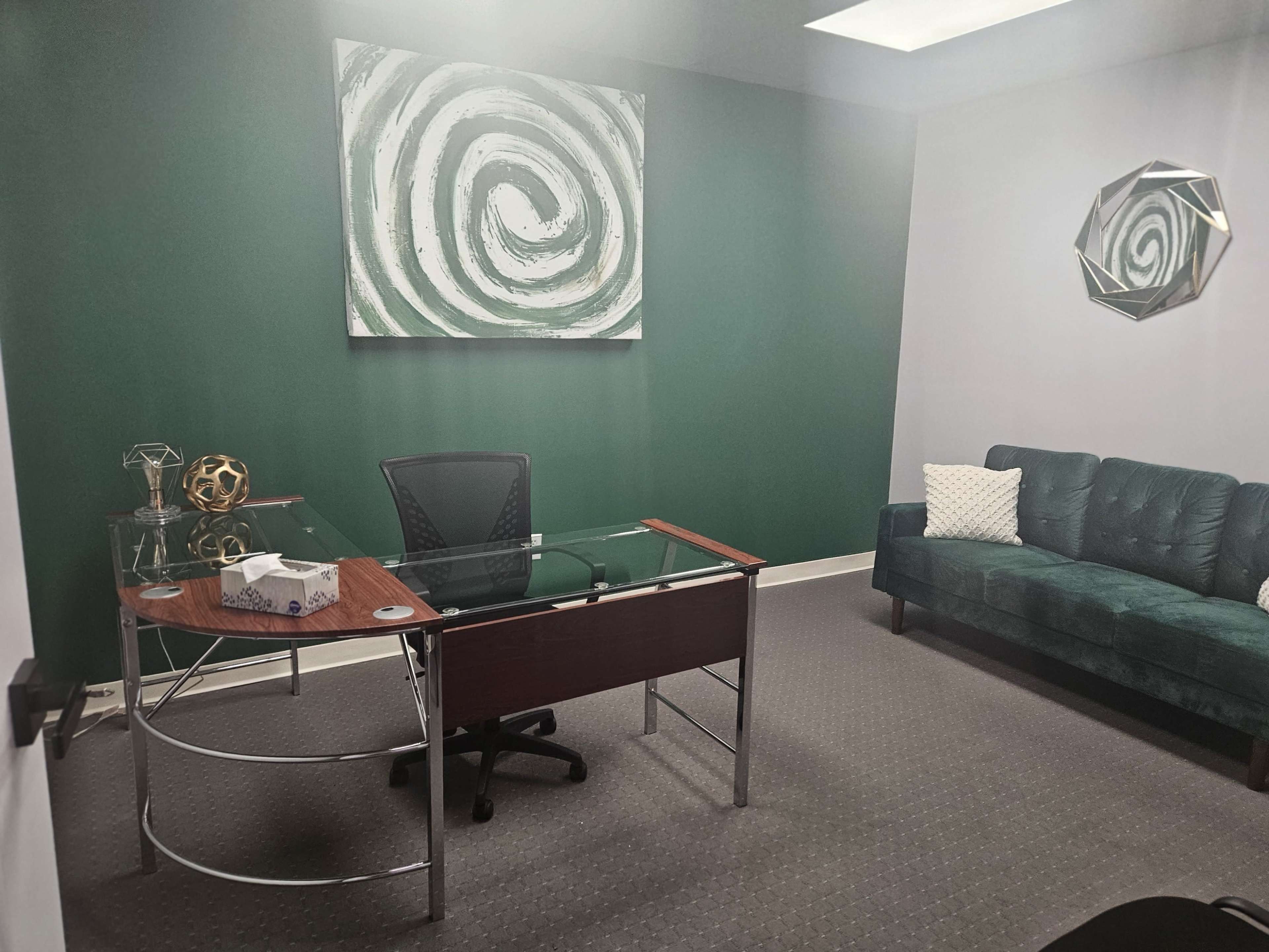 An office space featuring a glass-topped desk, a black office chair, a green accent wall with abstract art, and a green velvet couch.