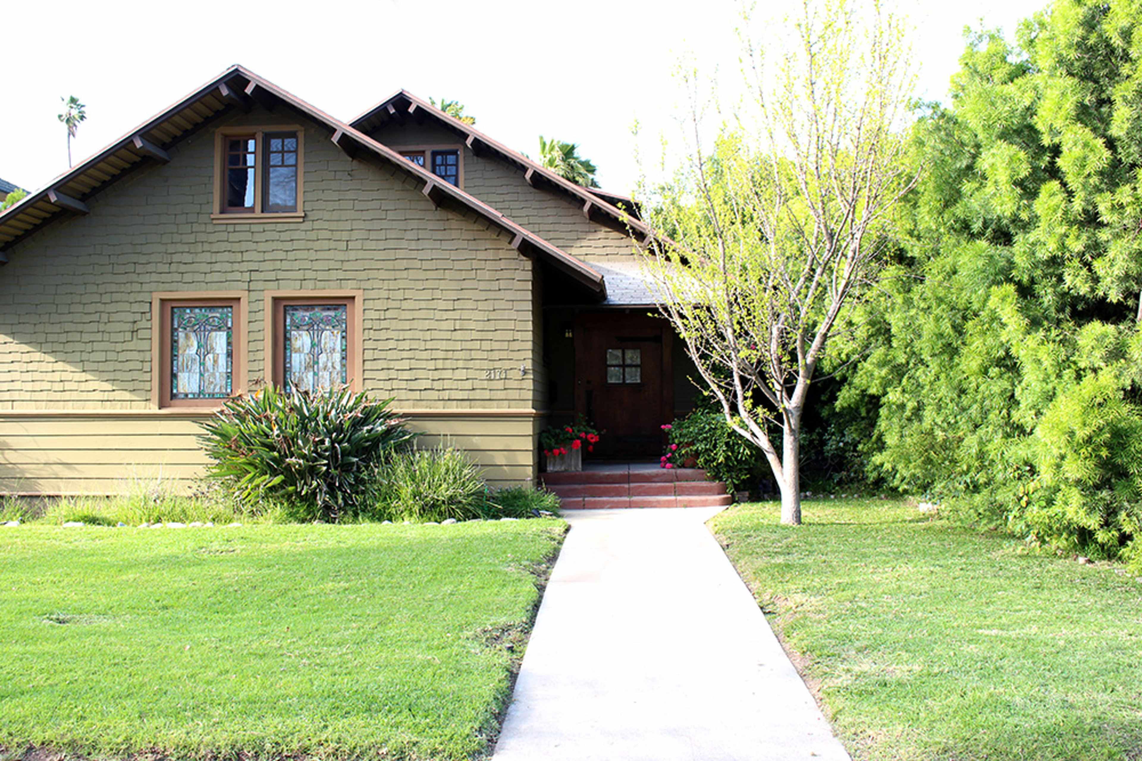 The image shows a single-story house with a front walkway, bordered by green grass and shrubs, and a small tree on the side.