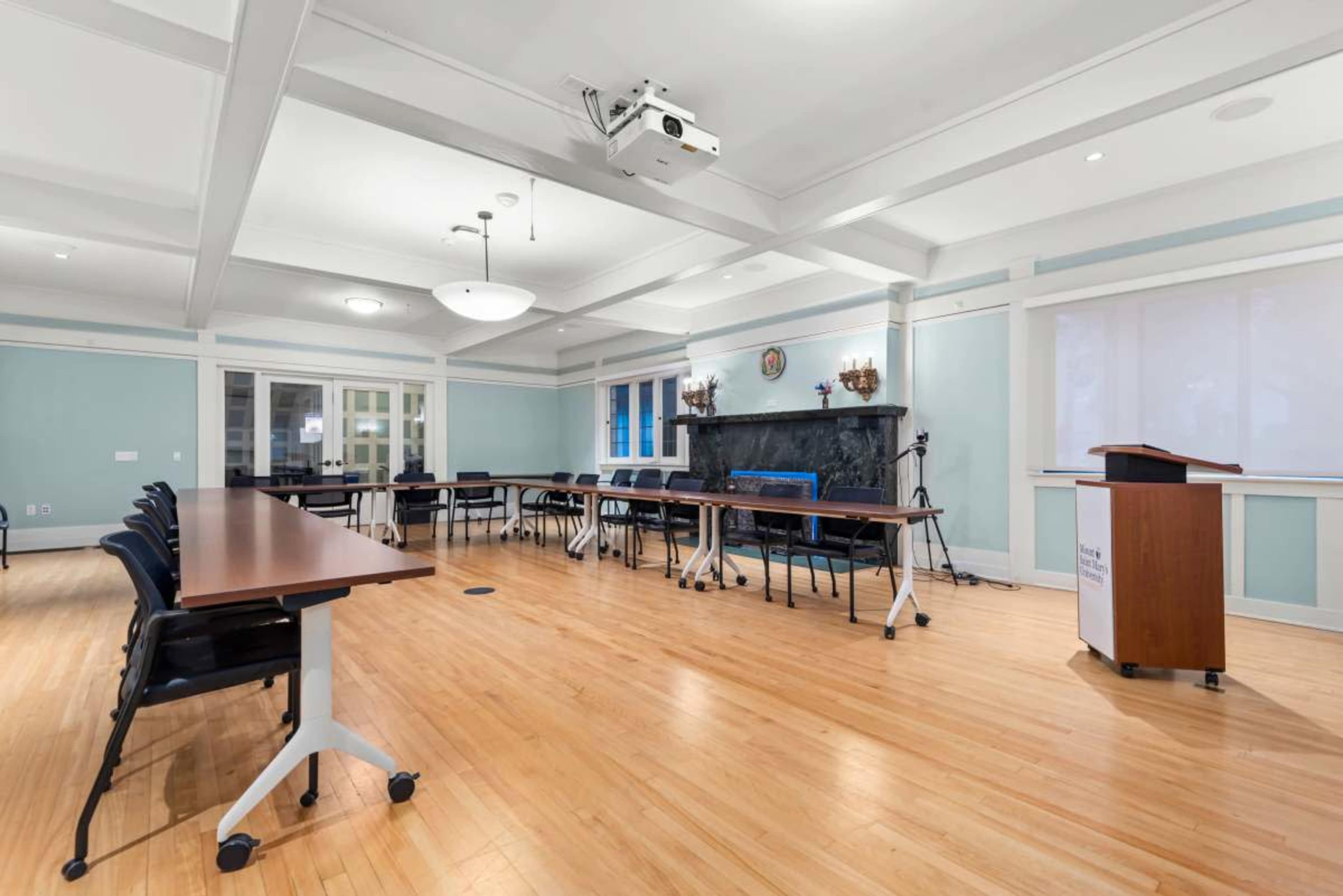 The image shows a well-lit meeting room with rows of tables and chairs arranged around a central space, featuring a podium, a projector, and a large window.