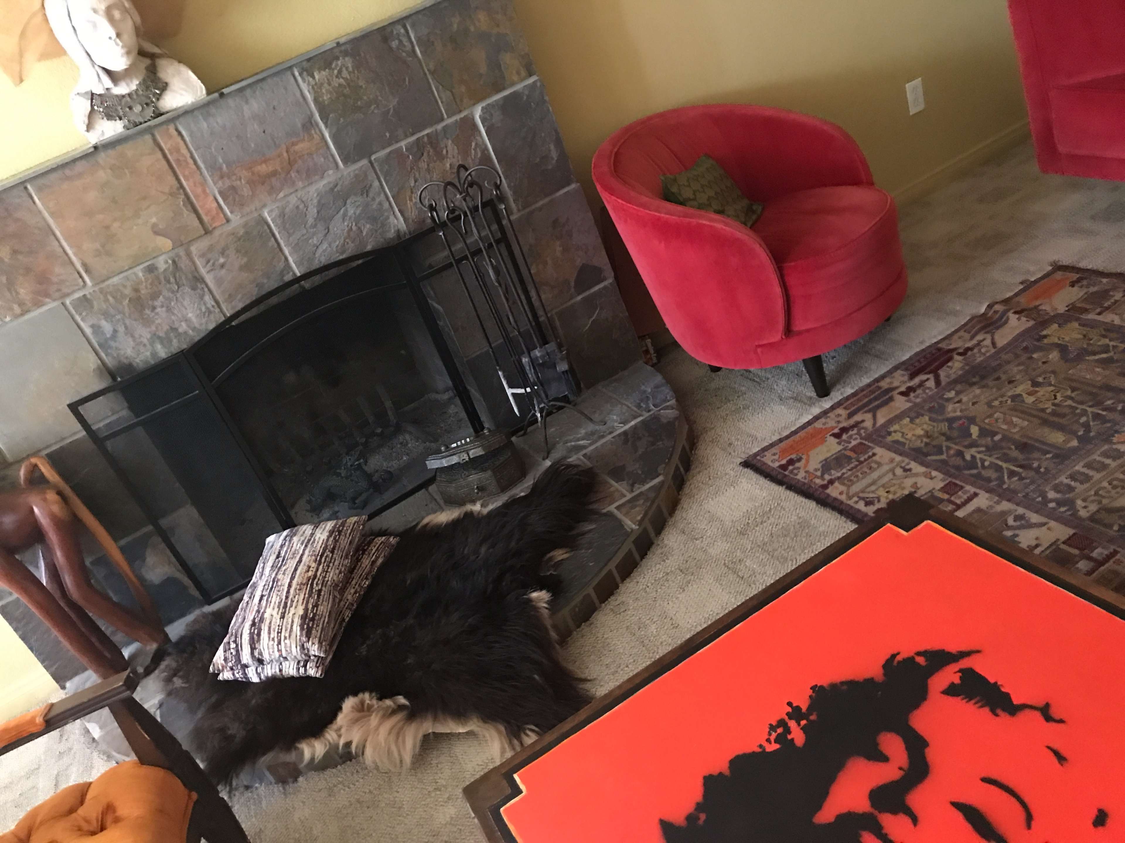 A living room with a stone fireplace, a red curved chair, a rug with intricate patterns, and a fur throw on the floor.