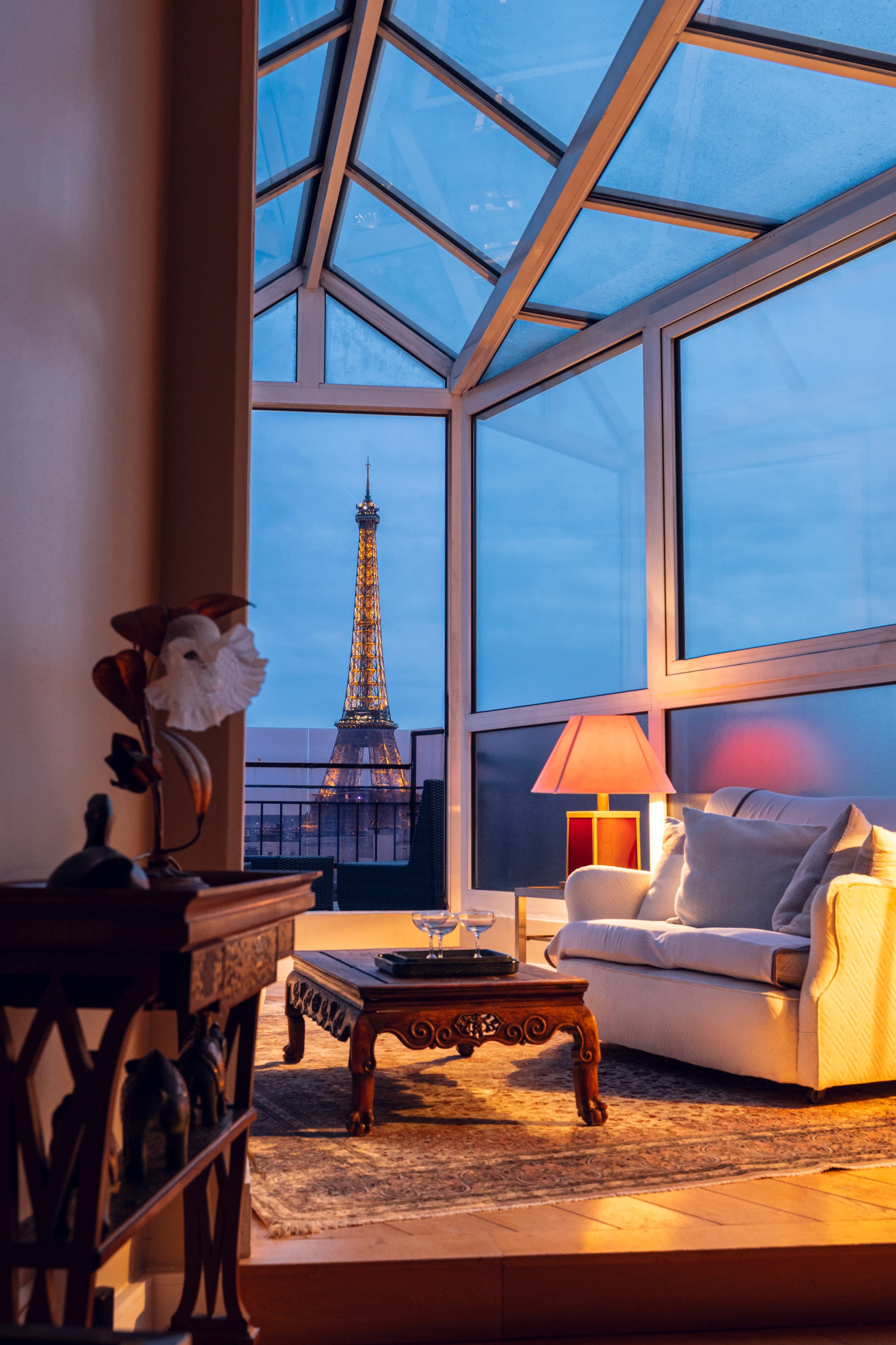 A cozy living room with glass walls offers a view of the Eiffel Tower illuminated at dusk.