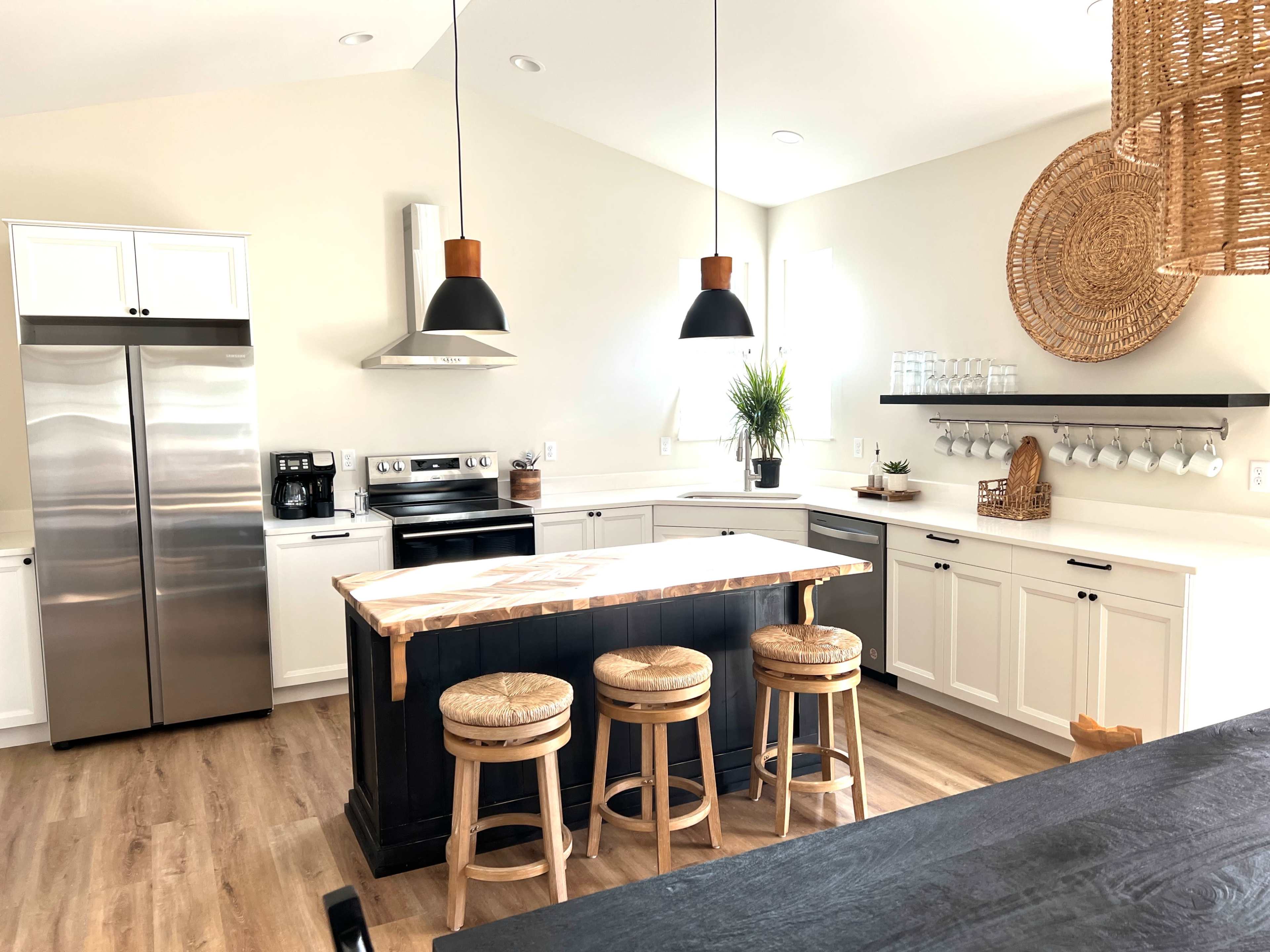 The image shows a modern kitchen featuring a central island with three wooden stools, stainless steel appliances, and a combination of white and dark cabinetry.