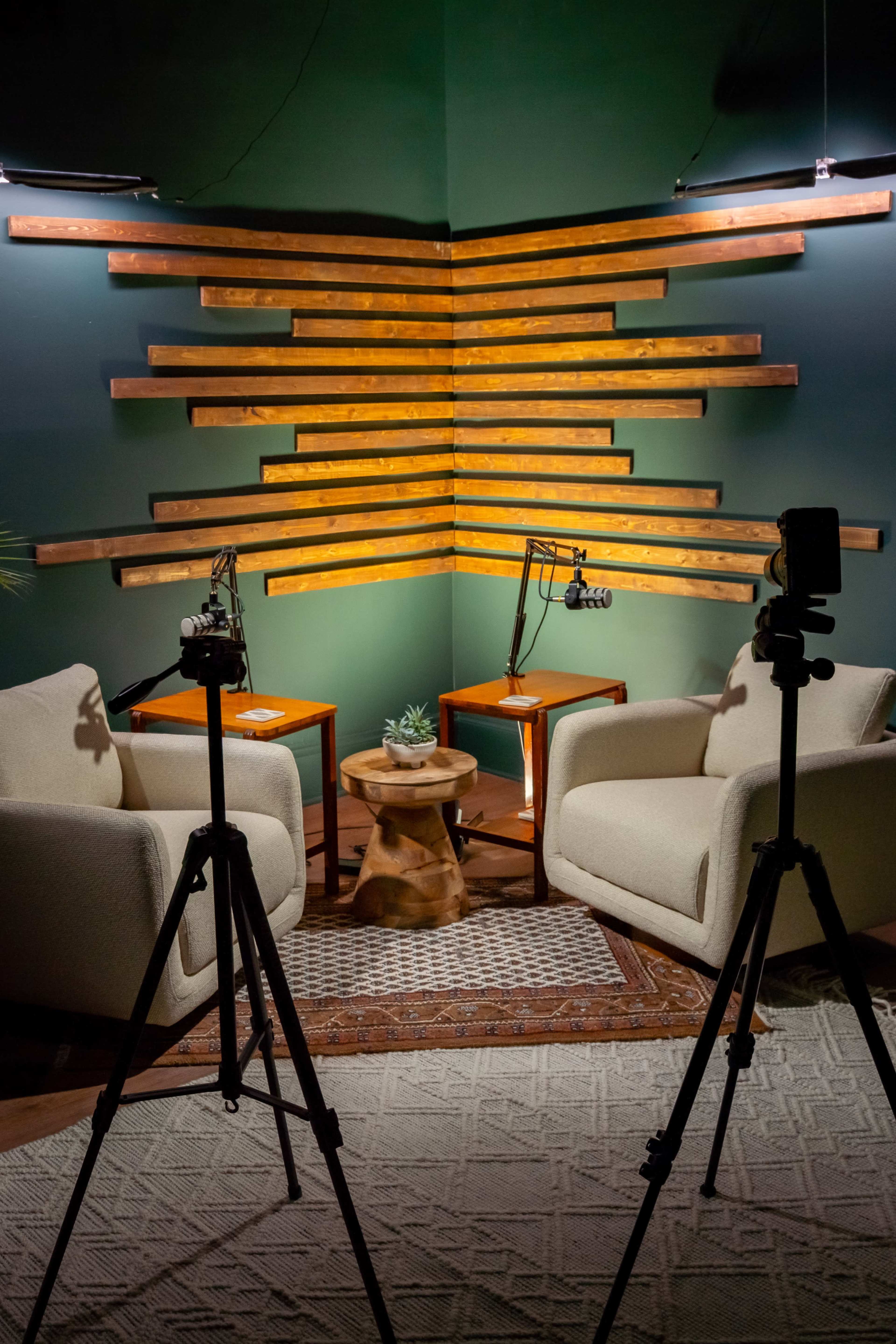 The image shows a cozy recording studio setup with two armchairs facing each other, separated by a small table, under warm lighting and a wooden slat wall backdrop.