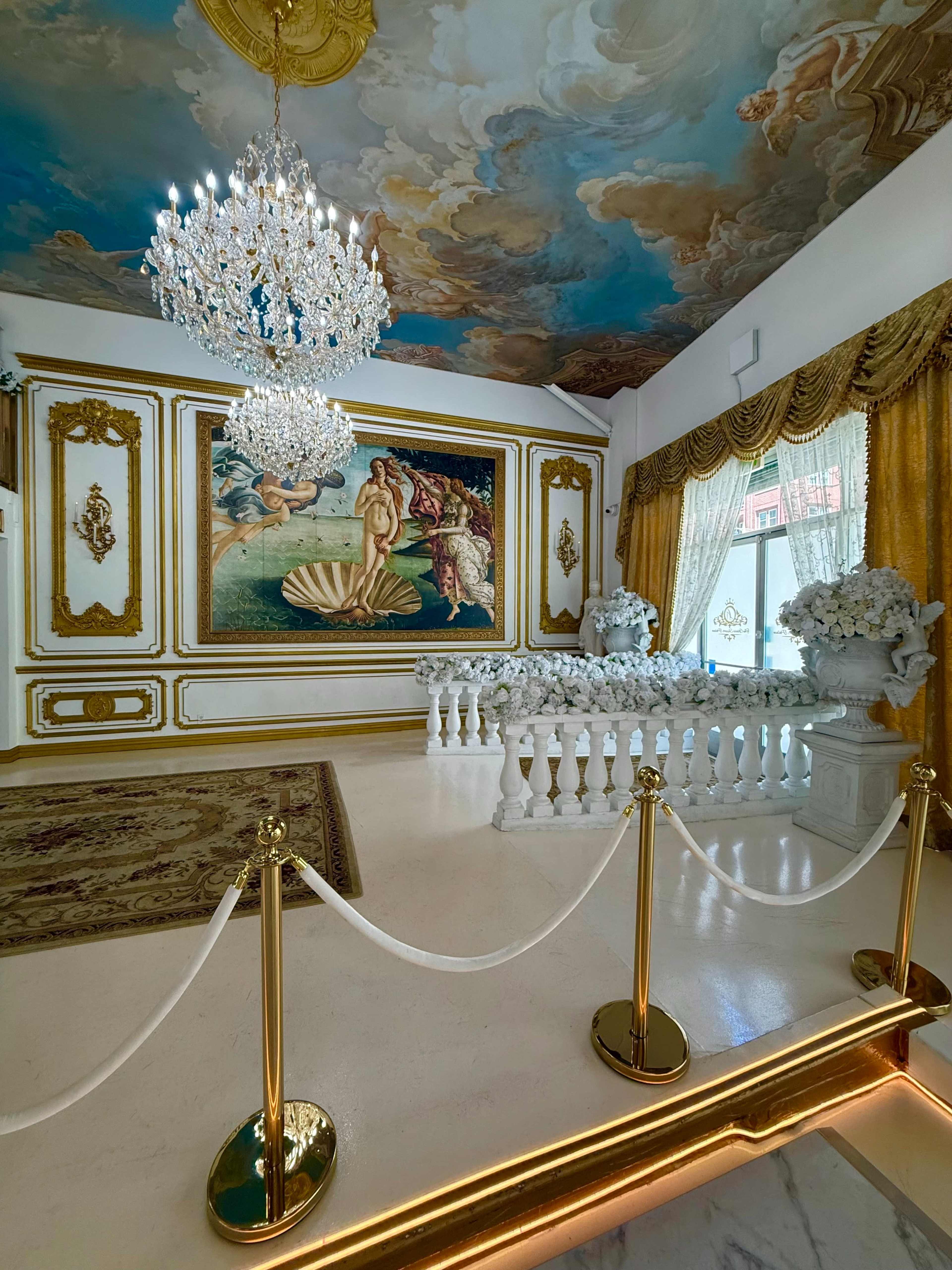 The image shows an opulent room featuring a painted ceiling, a large chandelier, a decorative mural on the wall, and elegant furnishings with floral arrangements.