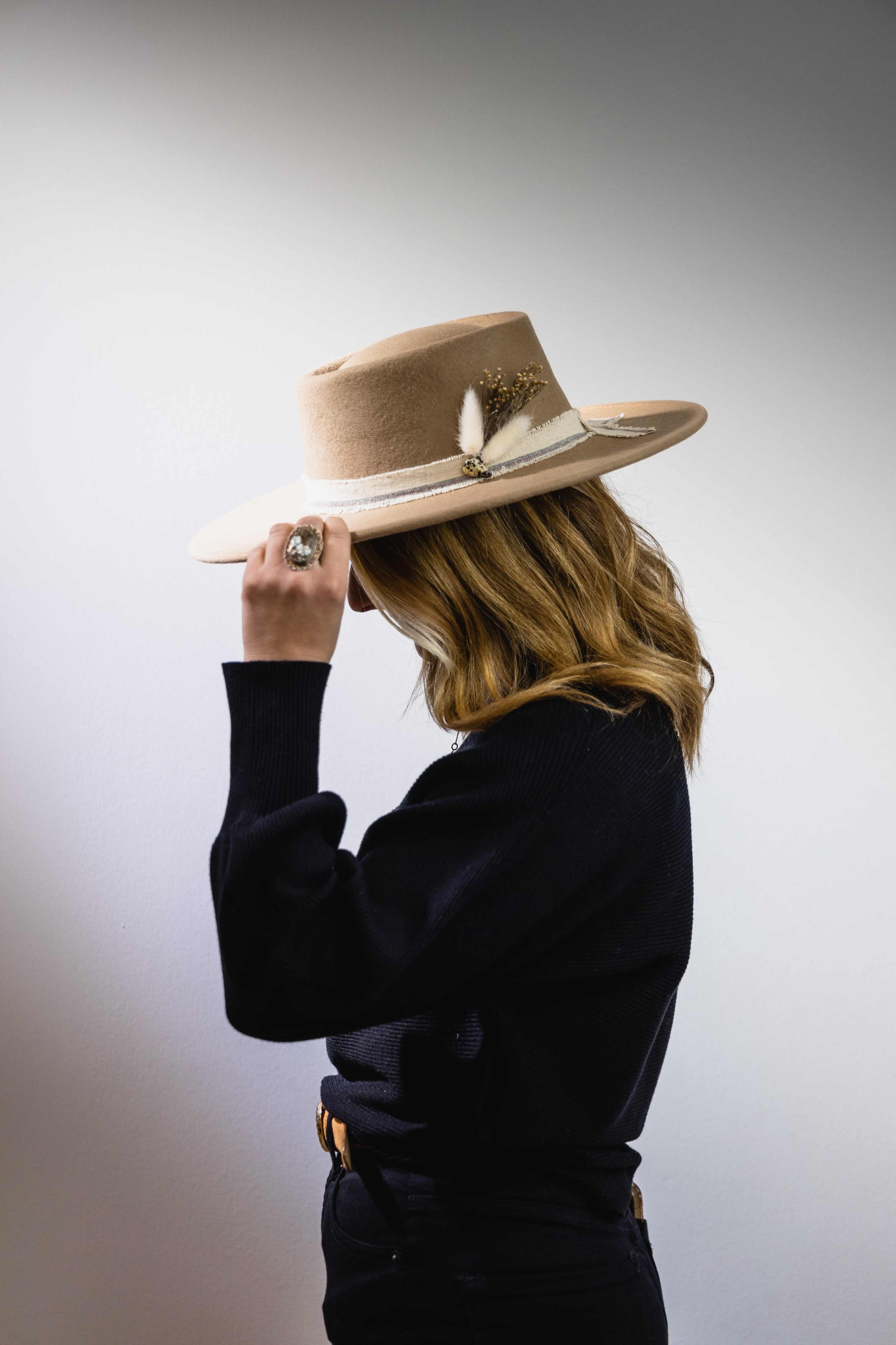 A person with shoulder-length hair is turning to the side while holding a wide-brimmed hat adorned with a feather.