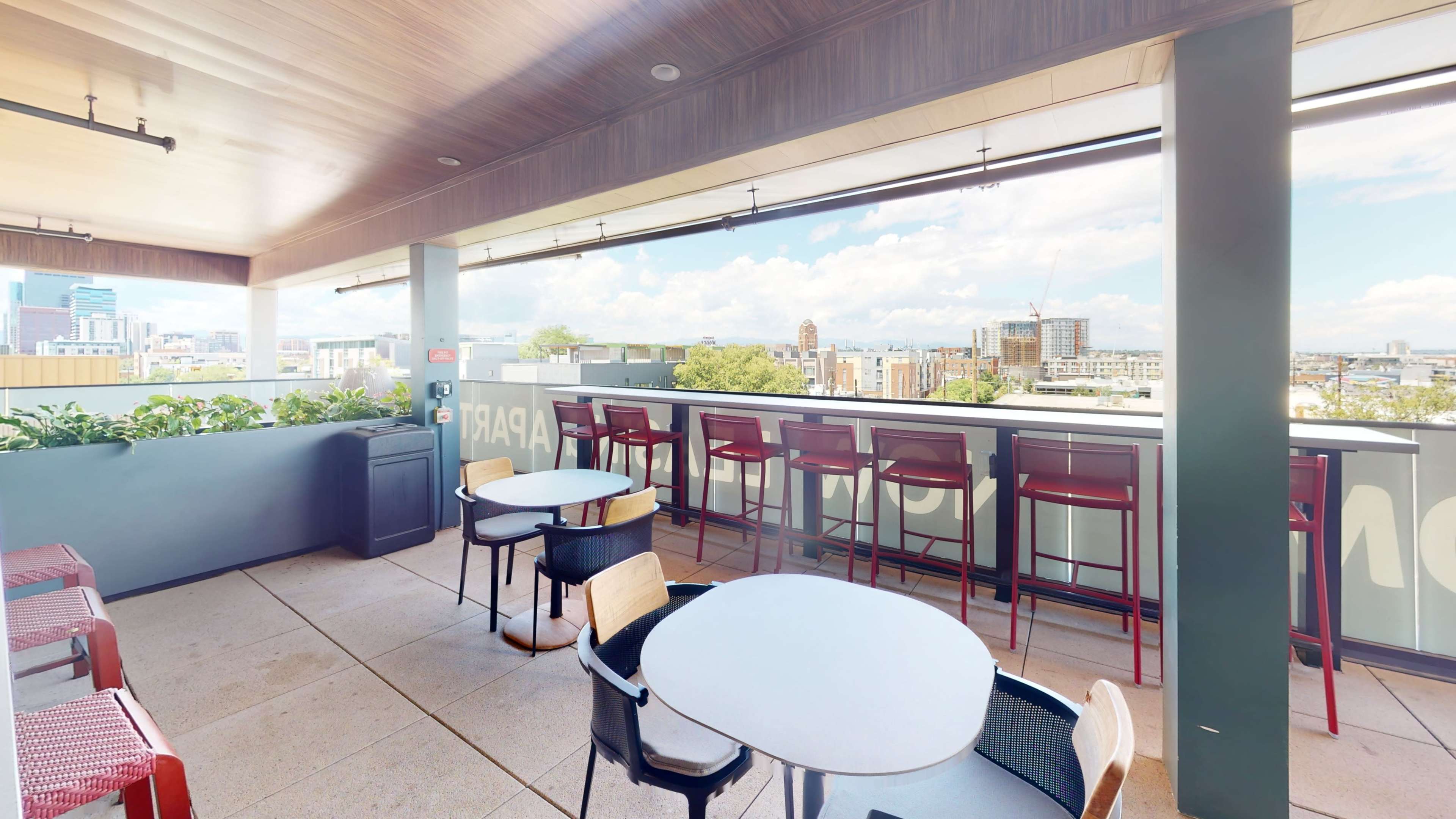 The image shows a rooftop terrace with several tables and chairs overlooking a city skyline.